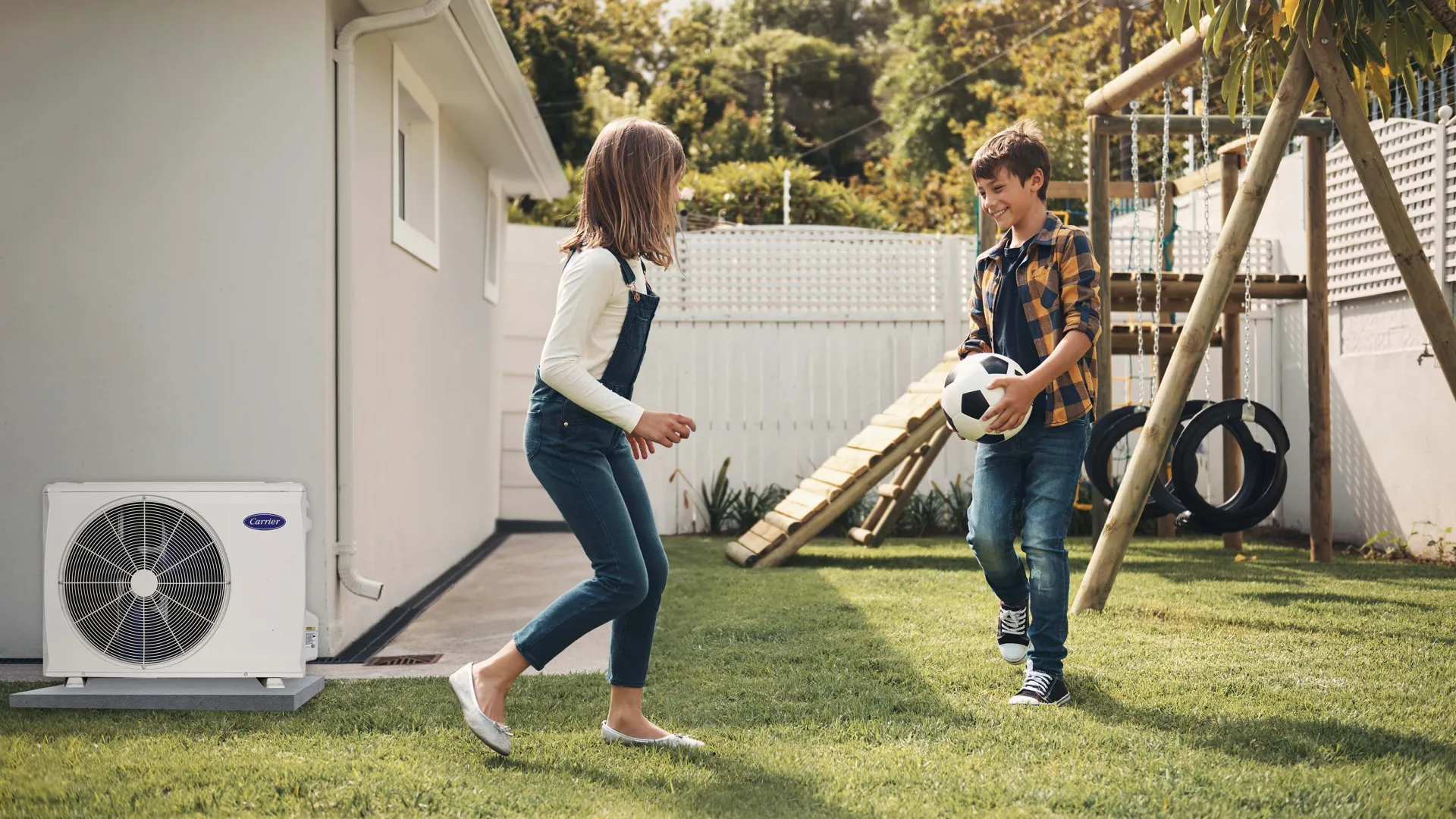 Two children playing with a soccer ball on a grassy backyard near a white house with an outdoor air conditioning unit.