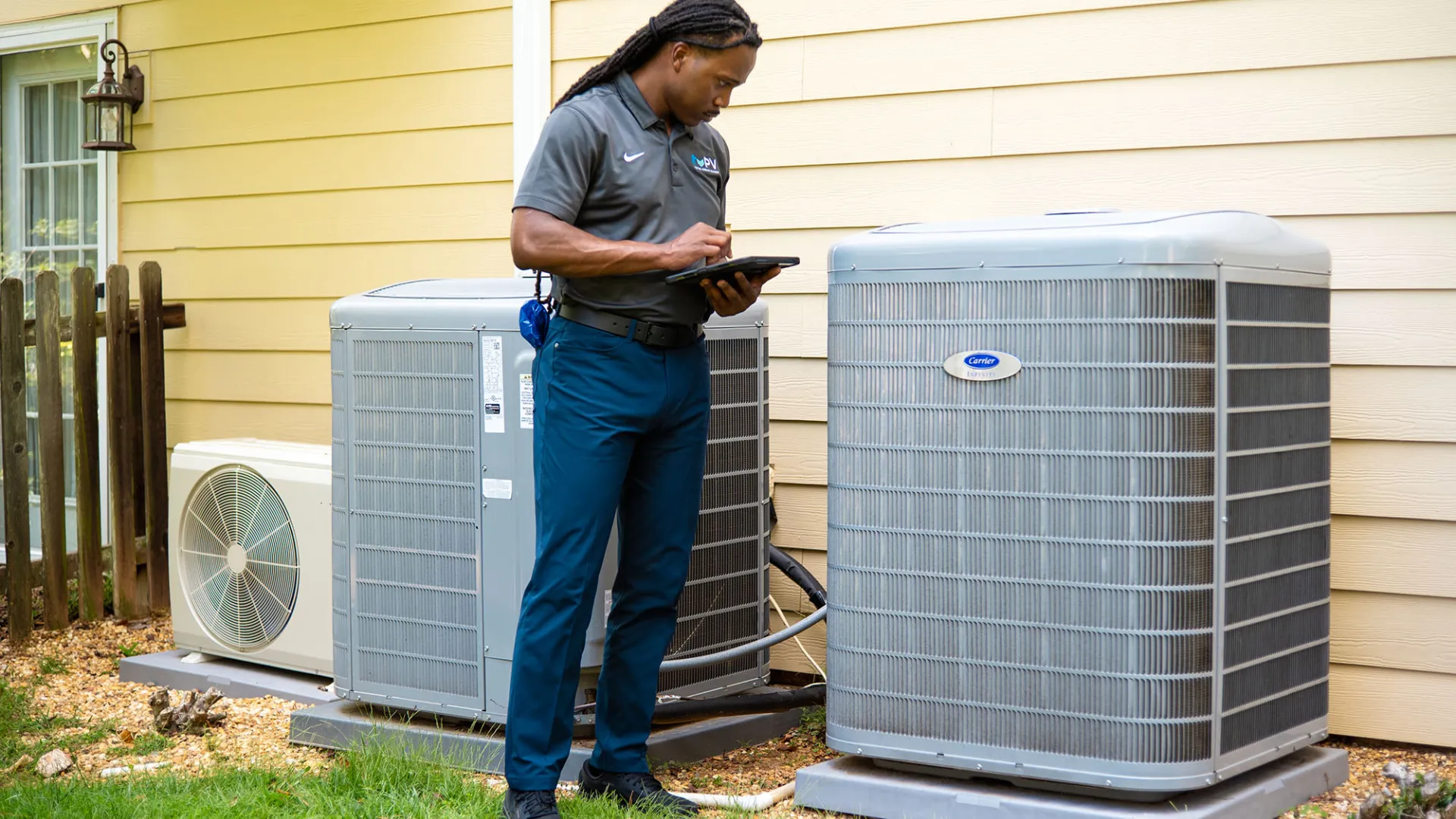 Technician inspecting and documenting the condition of three outdoor air conditioning units beside a house.