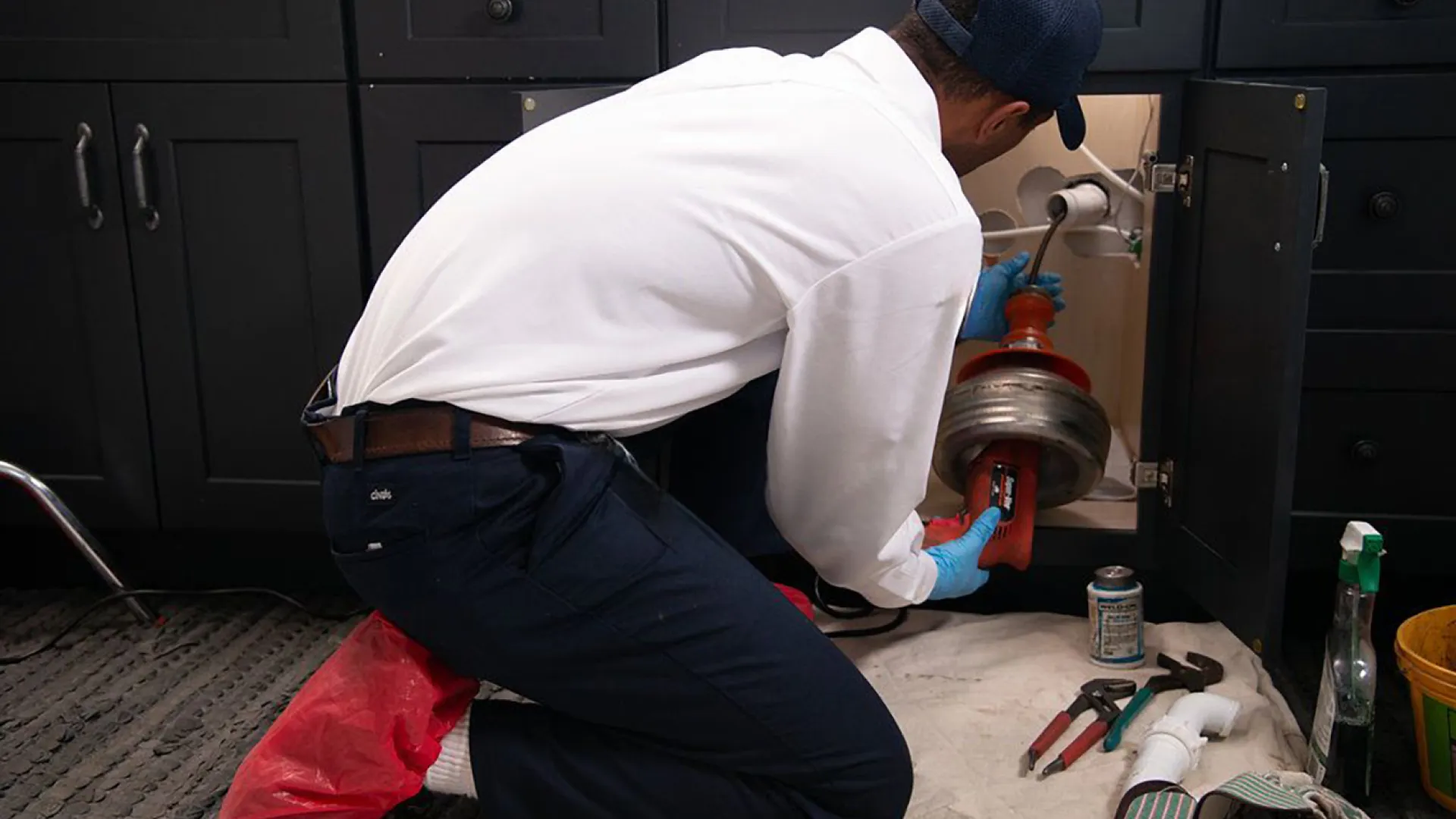 Plumber in gloves repairing kitchen sink drain with tools and cleaning supplies nearby on floor.