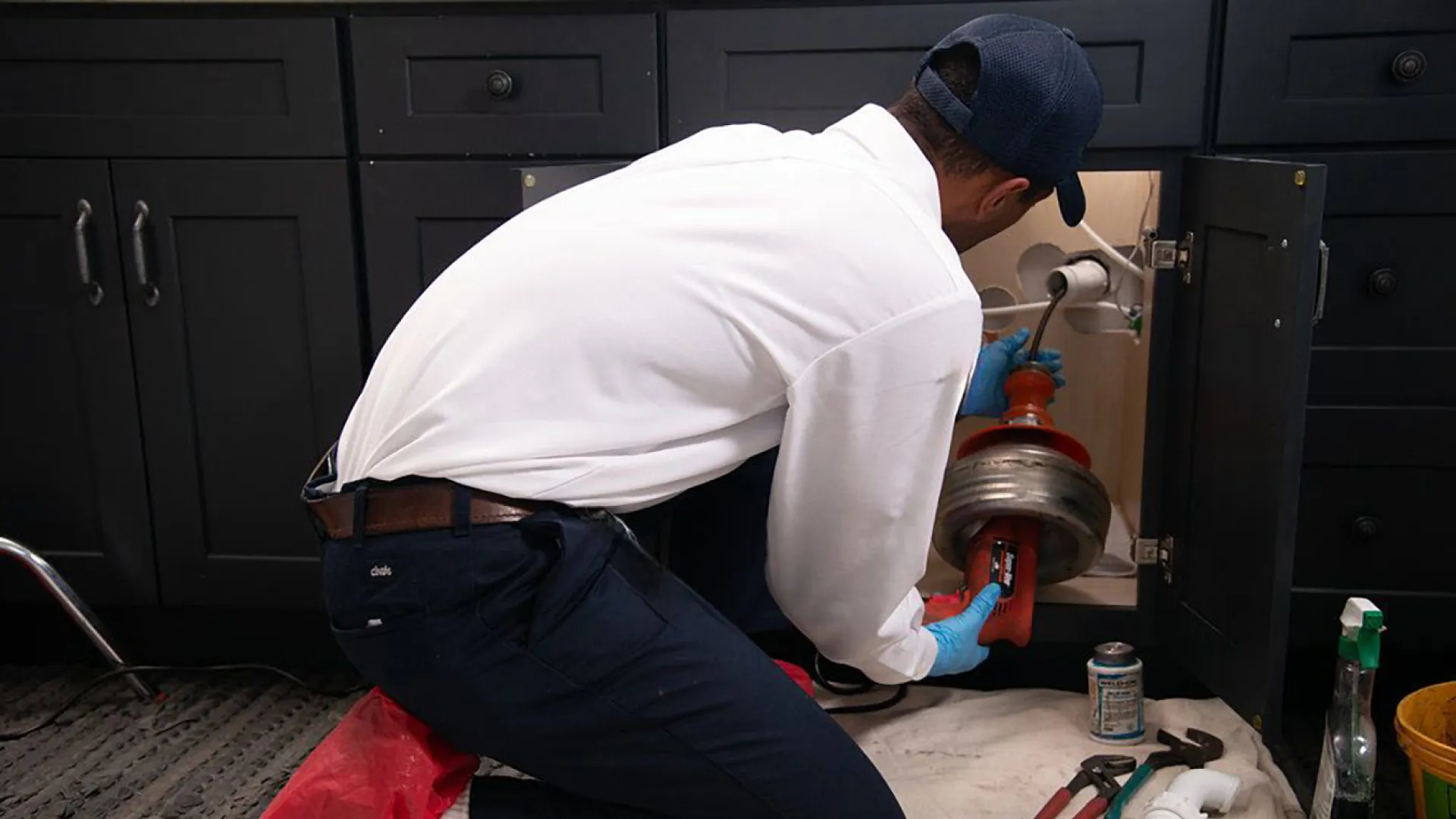 Plumber in gloves repairing kitchen sink drain with tools and cleaning supplies nearby on floor.