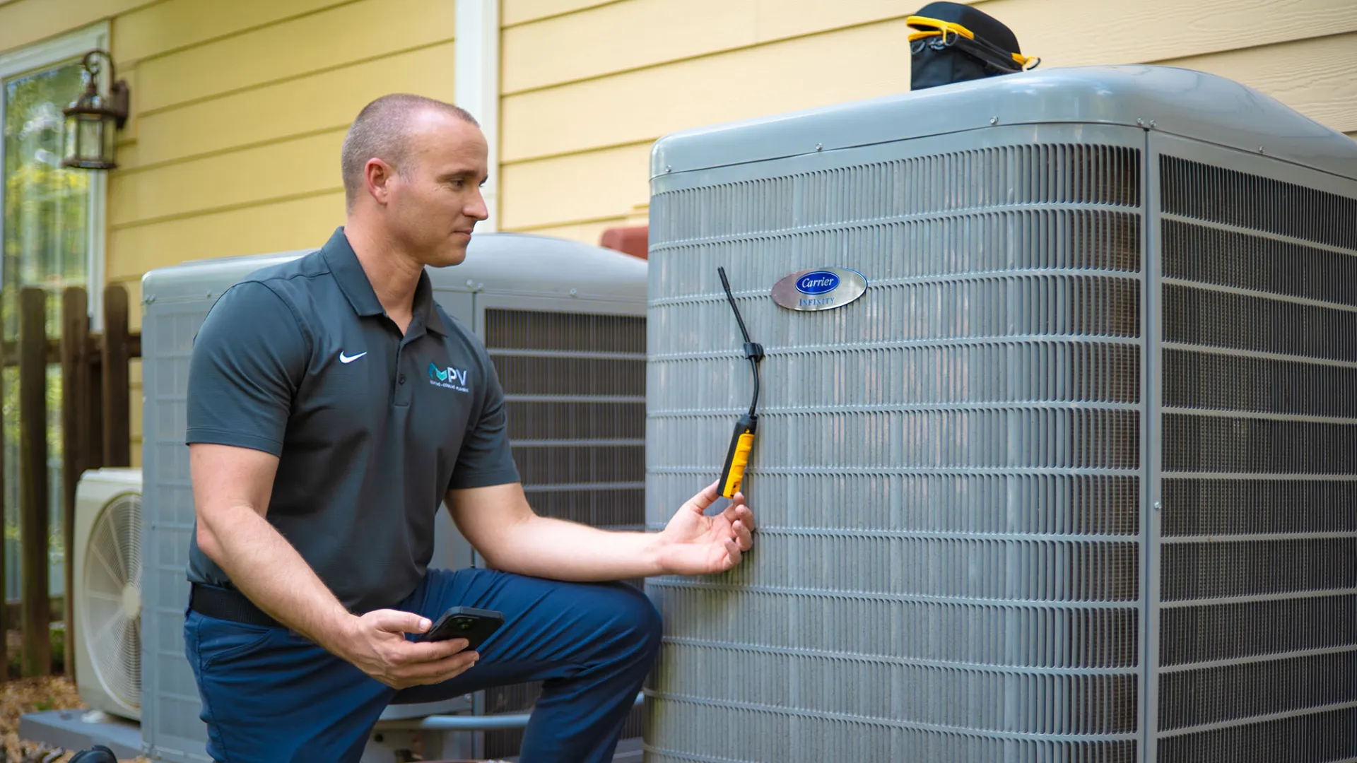 Technician inspecting an outdoor Carrier air conditioning unit using a diagnostic tool and smartphone in daylight.