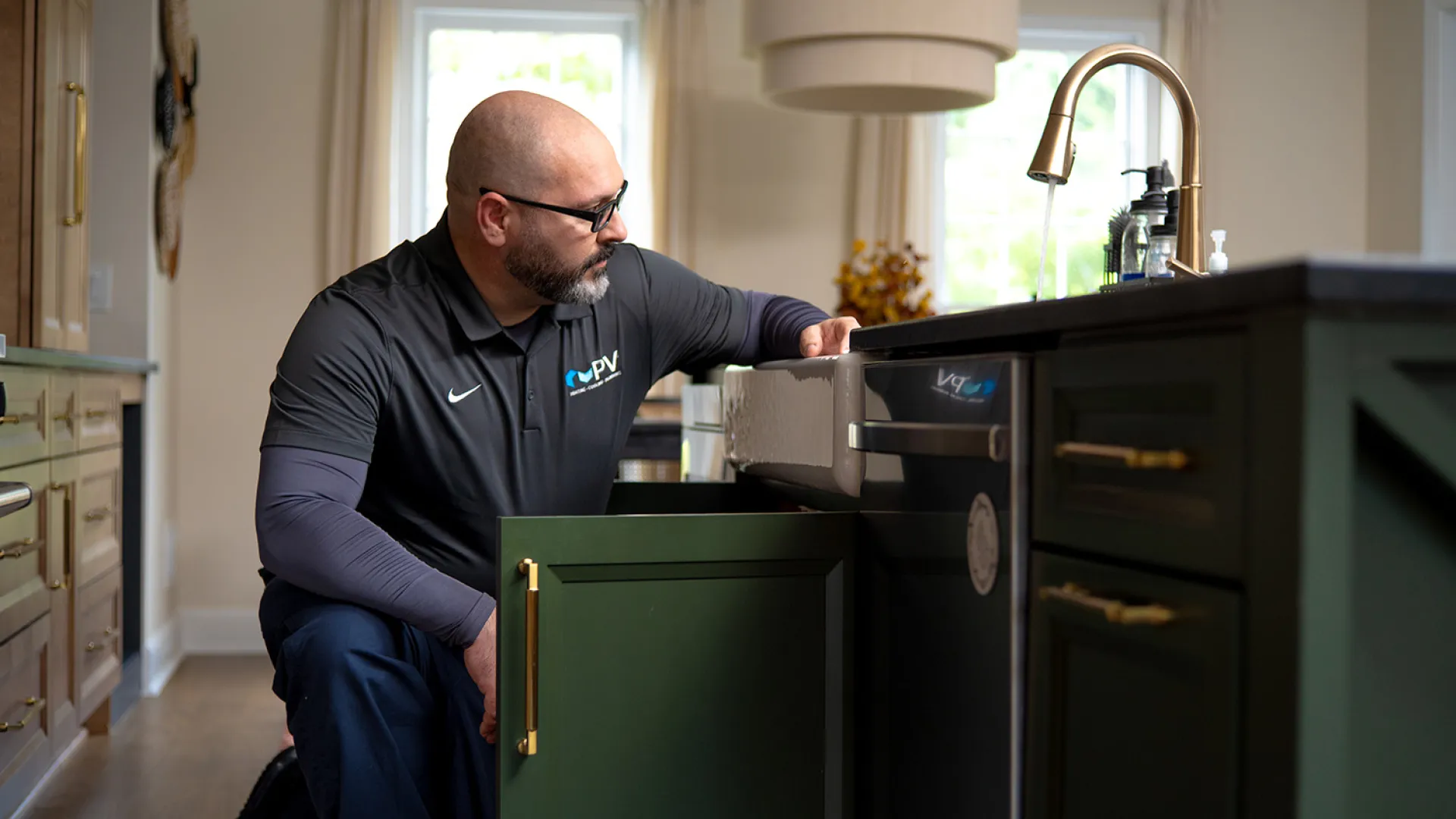 Technician inspecting plumbing under kitchen sink with green cabinets and gold handles in a modern home.