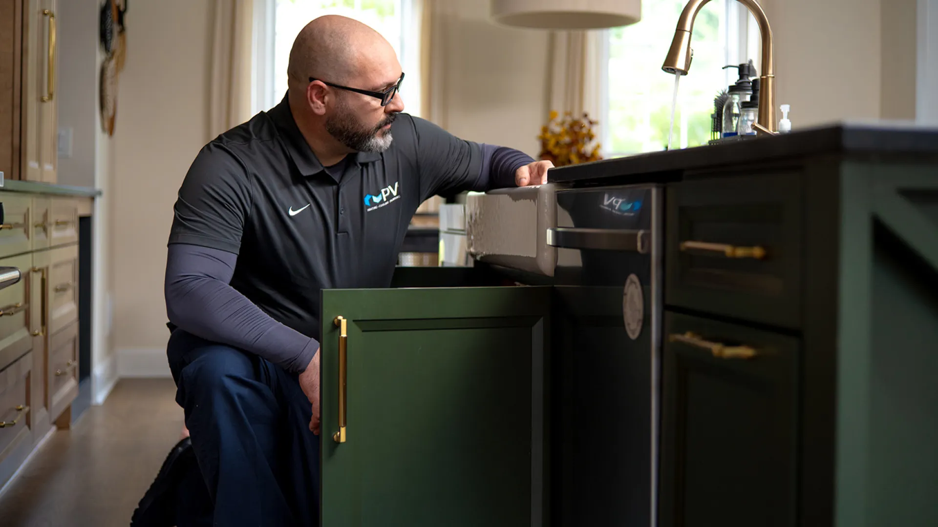 Technician inspecting plumbing under kitchen sink with green cabinets and gold handles in a modern home.
