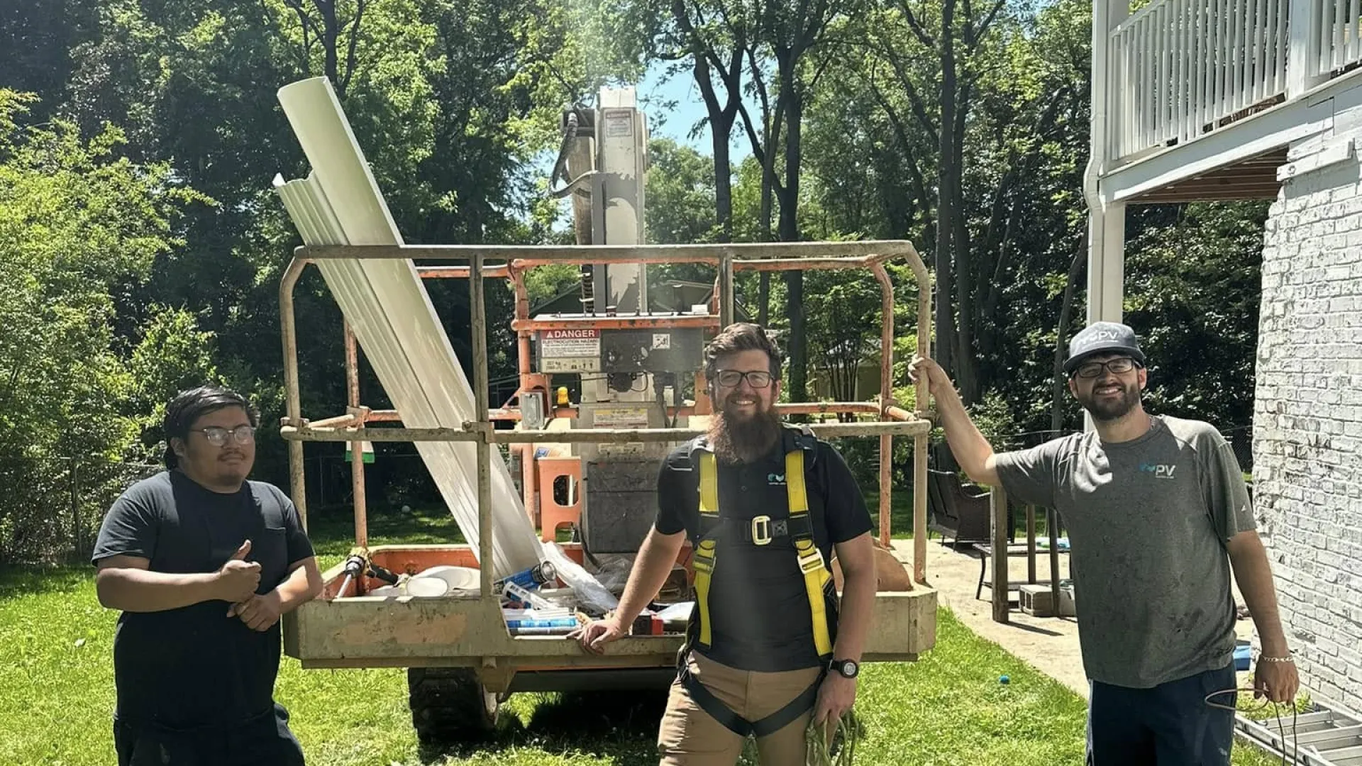 Three construction workers standing on grass next to equipment and a house on a sunny day