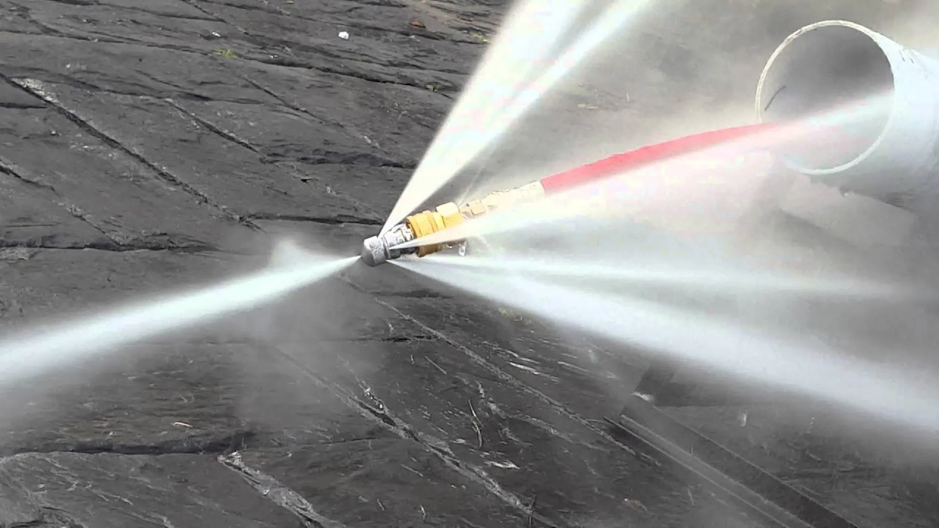 High-pressure water jet nozzle spraying multiple streams for surface cleaning on dark stone pavement.