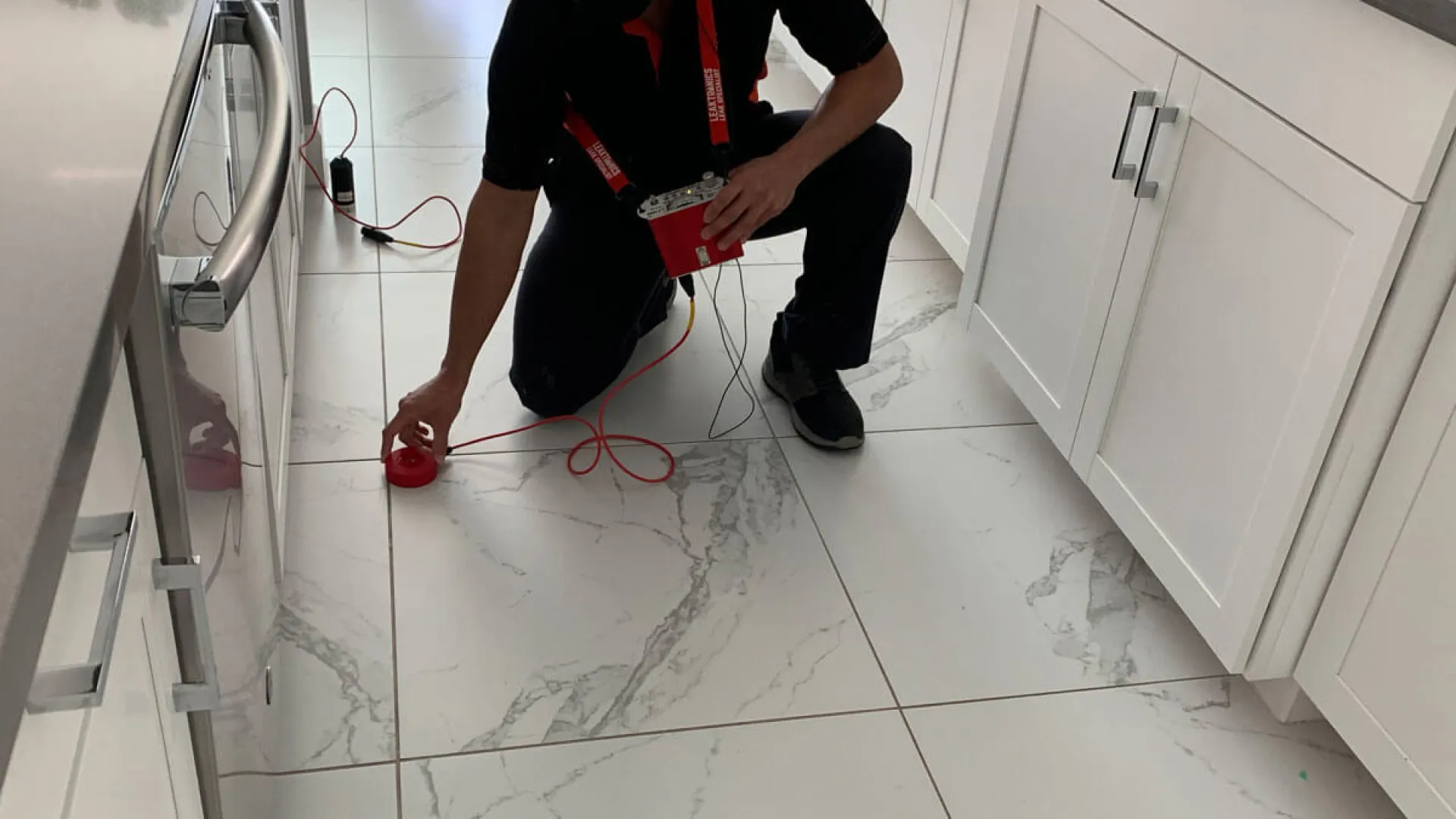 Technician in black uniform inspecting kitchen floor with red testing device and wiring in modern white kitchen.