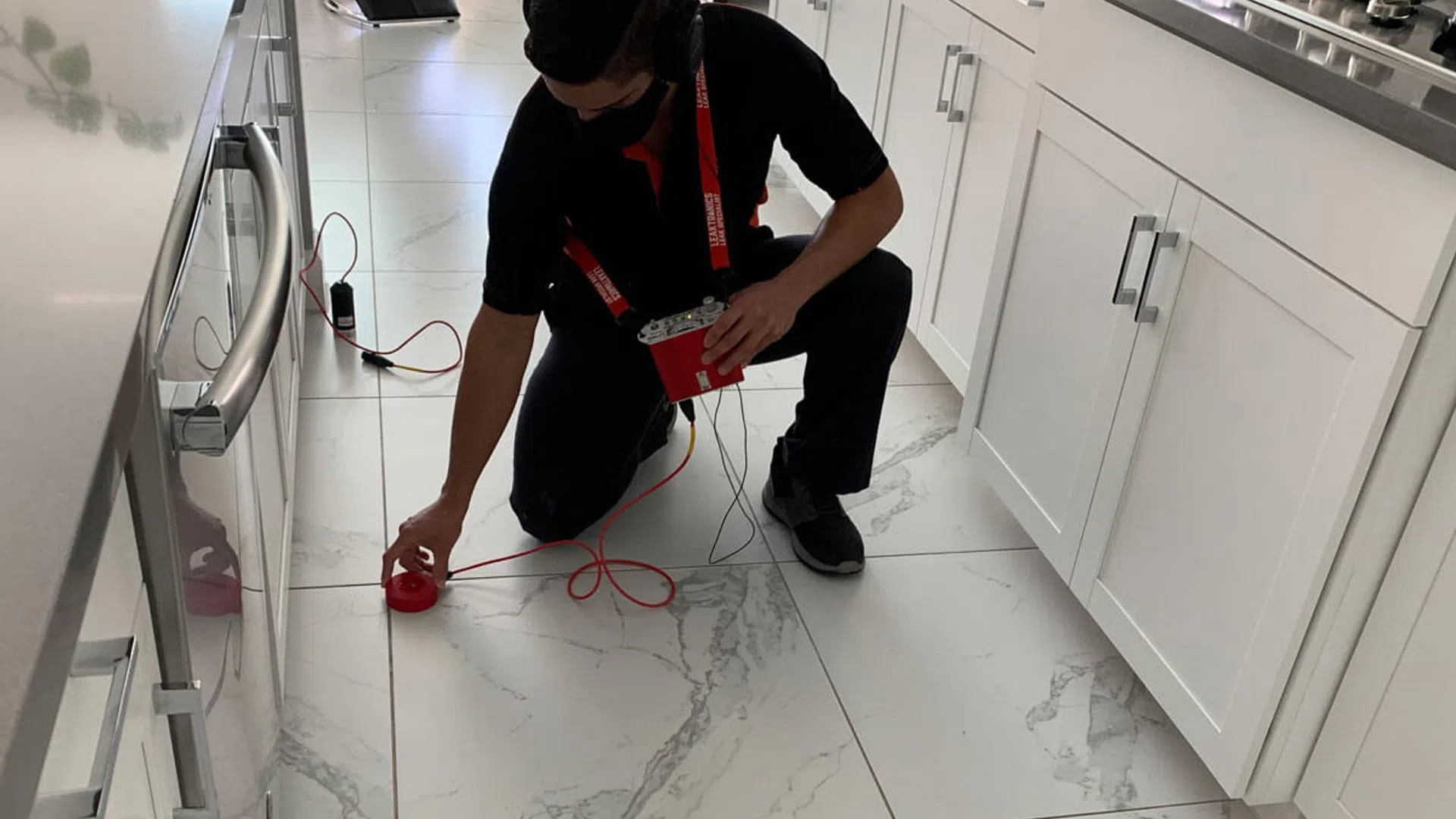 Technician with headphones using electronic equipment to inspect white kitchen floor tiles by cabinets and stove.