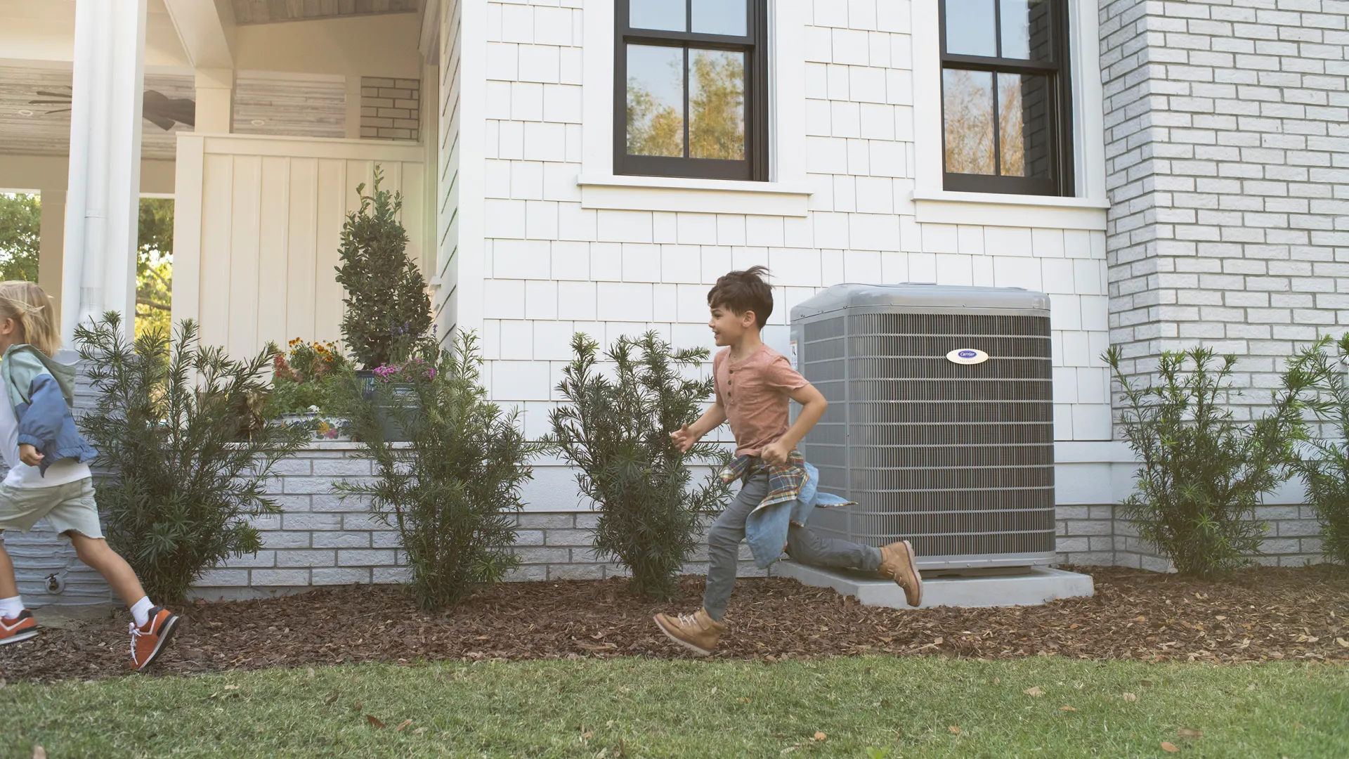 Two children running and playing near a white house with an outdoor air conditioning unit on a sunny day