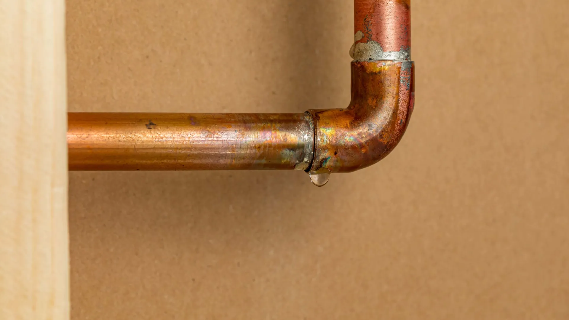 Close-up of a leaking copper pipe joint with a water droplet against a brown background.