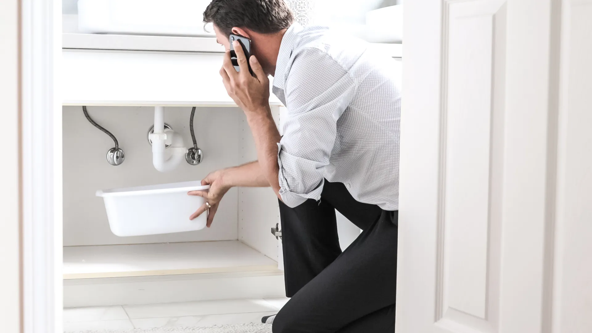 Man troubleshooting a sink issue while on the phone, kneeling beside a bathroom vanity cabinet.
