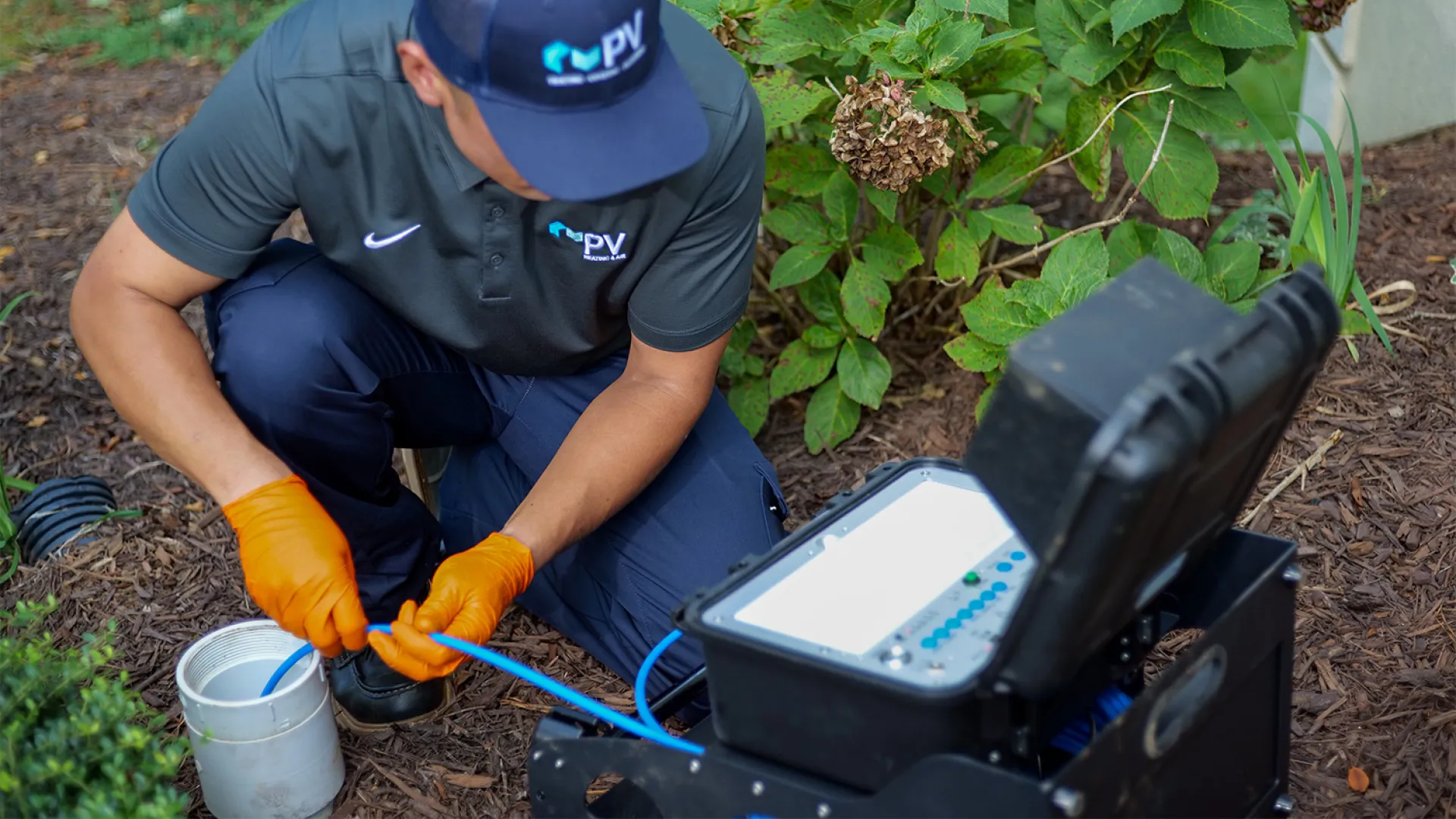 Technician in blue uniform and orange gloves working with fiber optic cables near a house garden.