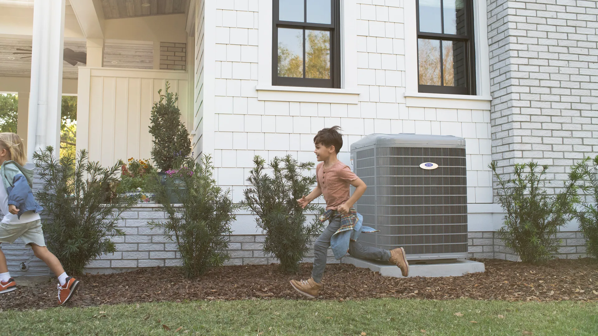 Two children running and playing outside a white house with an air conditioning unit against the wall