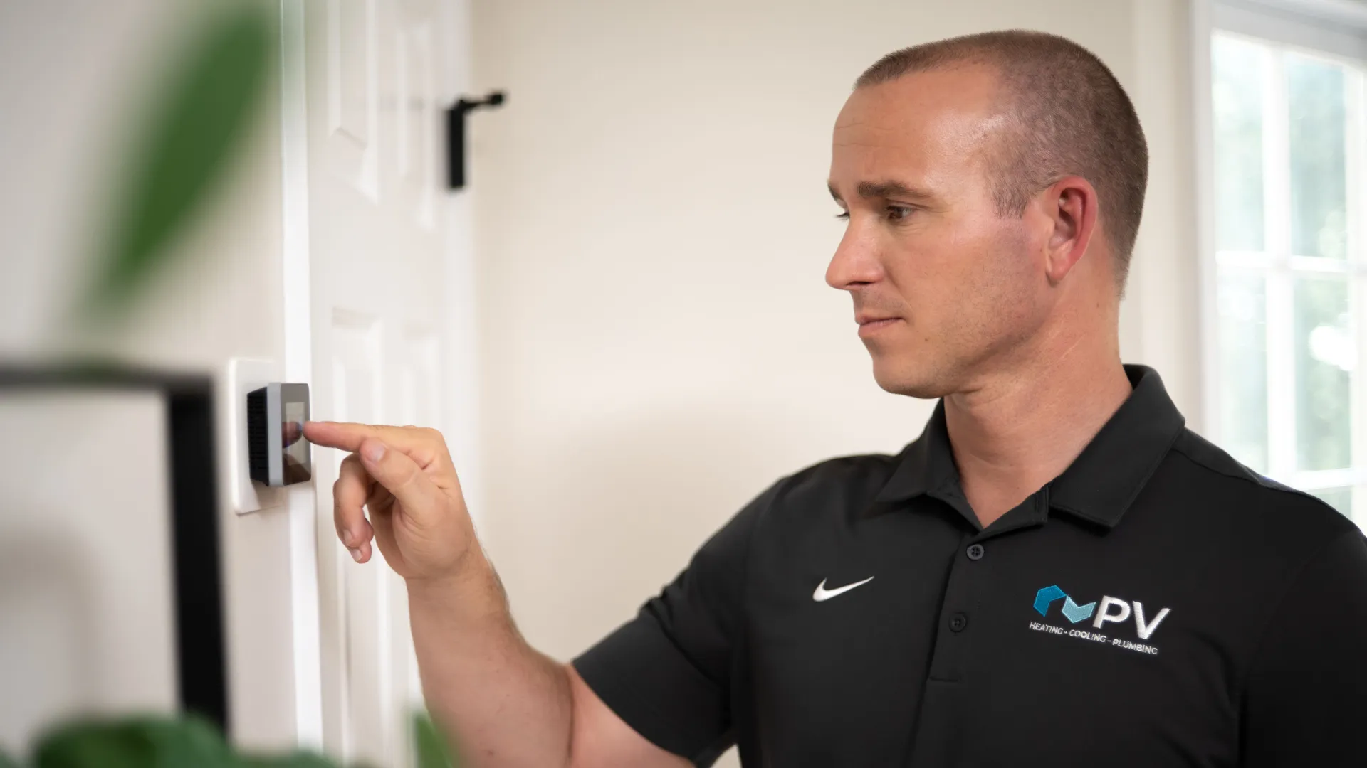 Man in black polo shirt adjusting digital thermostat on white wall in bright room with natural light.