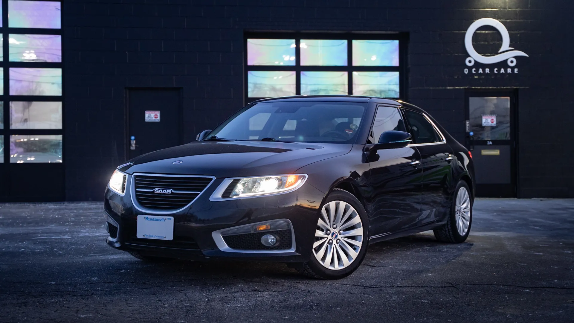 Black Saab sedan parked outside a dark building with colorful windows at dusk and headlights on.