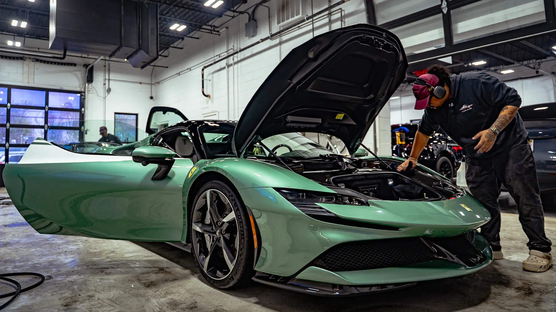 Green sports car with open hood and door being inspected by a mechanic in a garage with bright lighting.