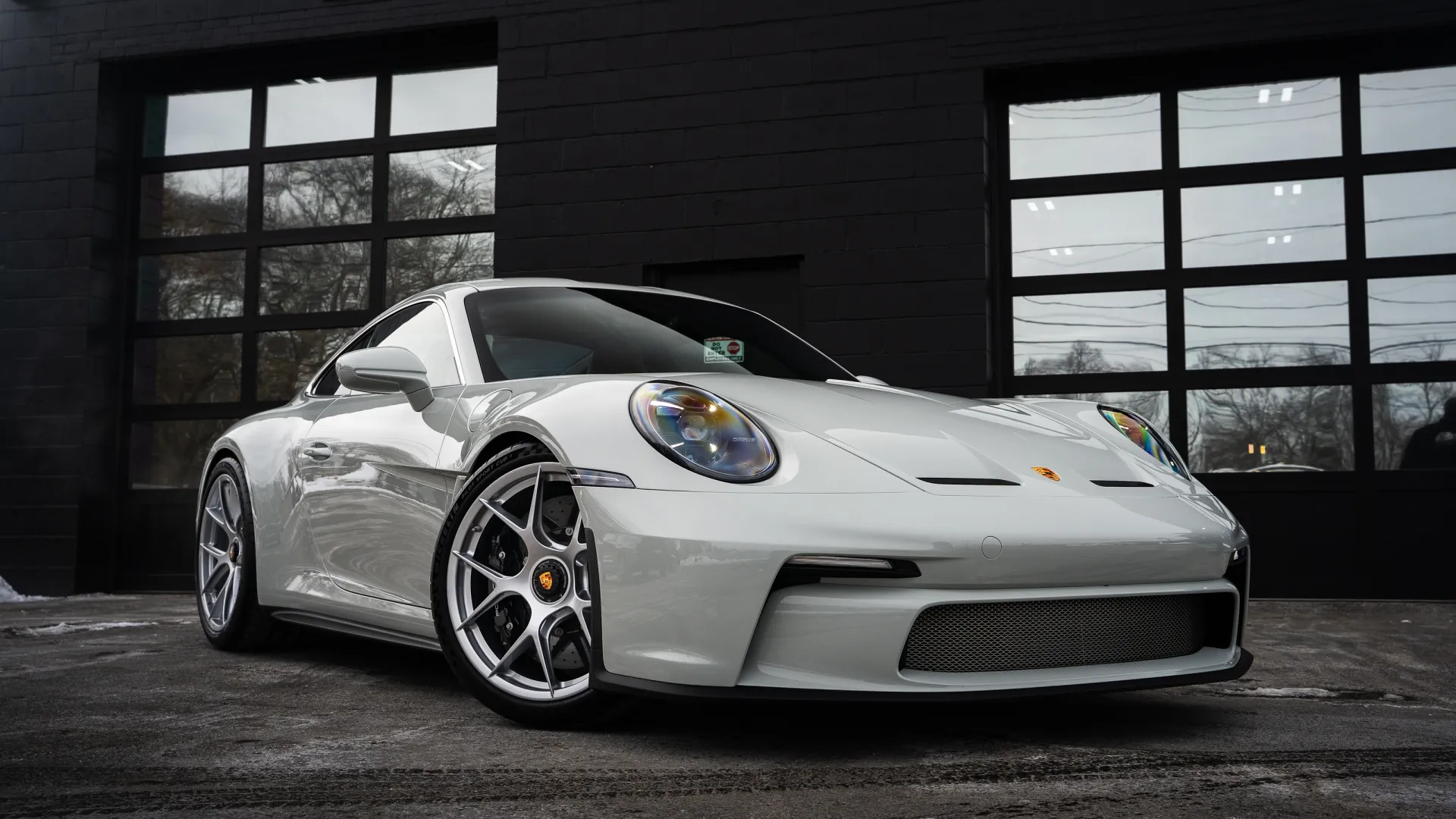 White Porsche sports car parked on asphalt in front of modern glass garage doors with reflections.