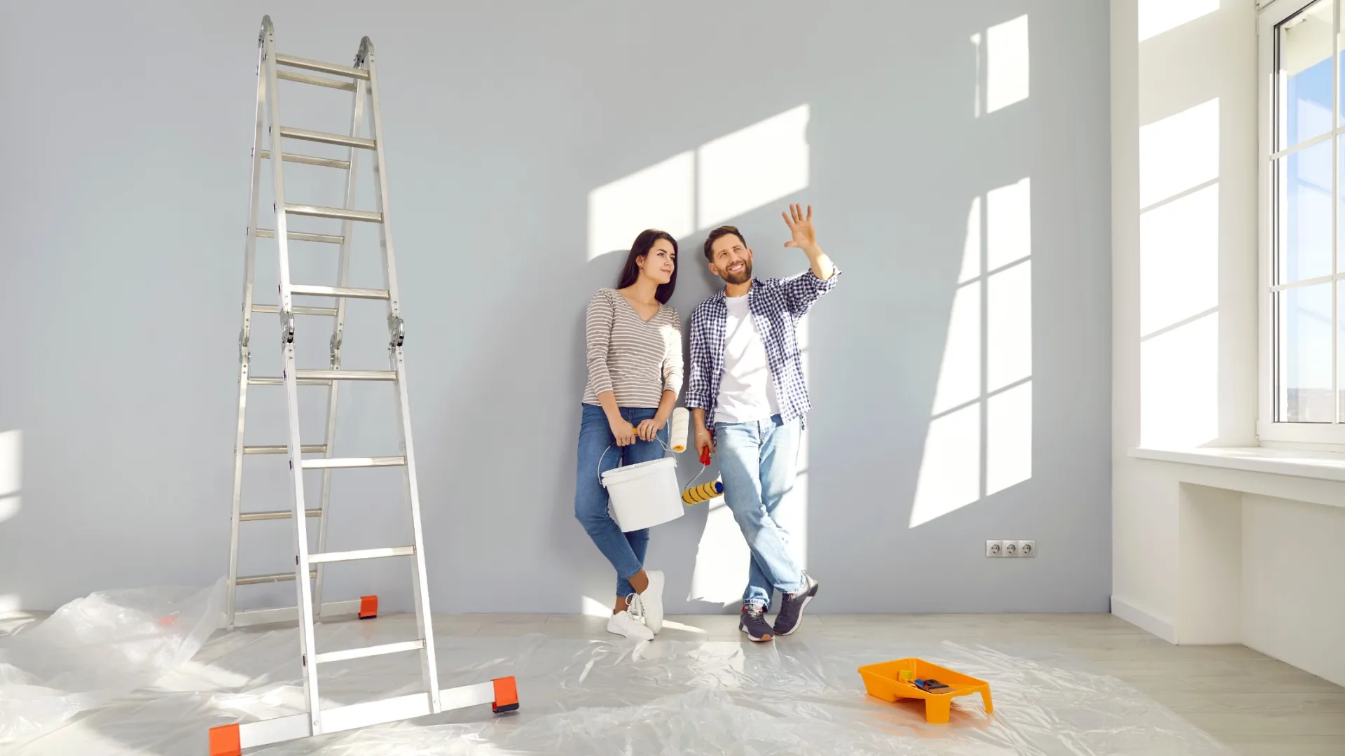 Couple leaning against wall in bright room with ladder and painting tools, planning home renovation project.