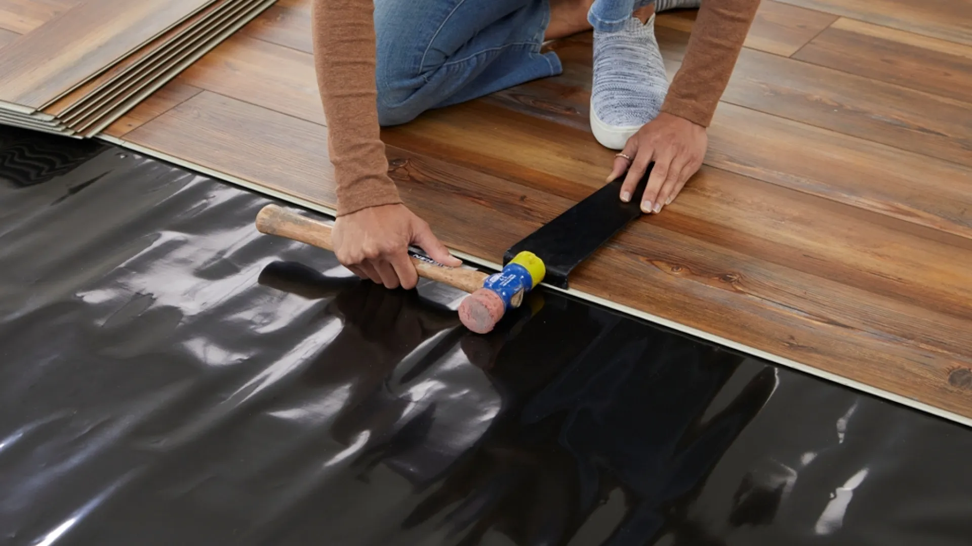 Person installing wood laminate flooring using a rubber mallet and tapping block on black underlayment.