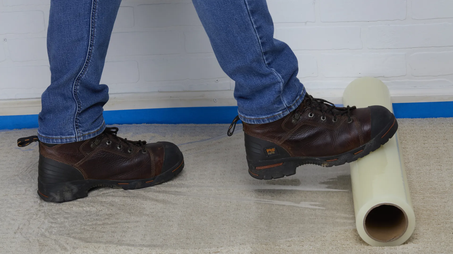 Person wearing brown boots stepping on a beige roll of material on a carpeted floor against a white brick wall.