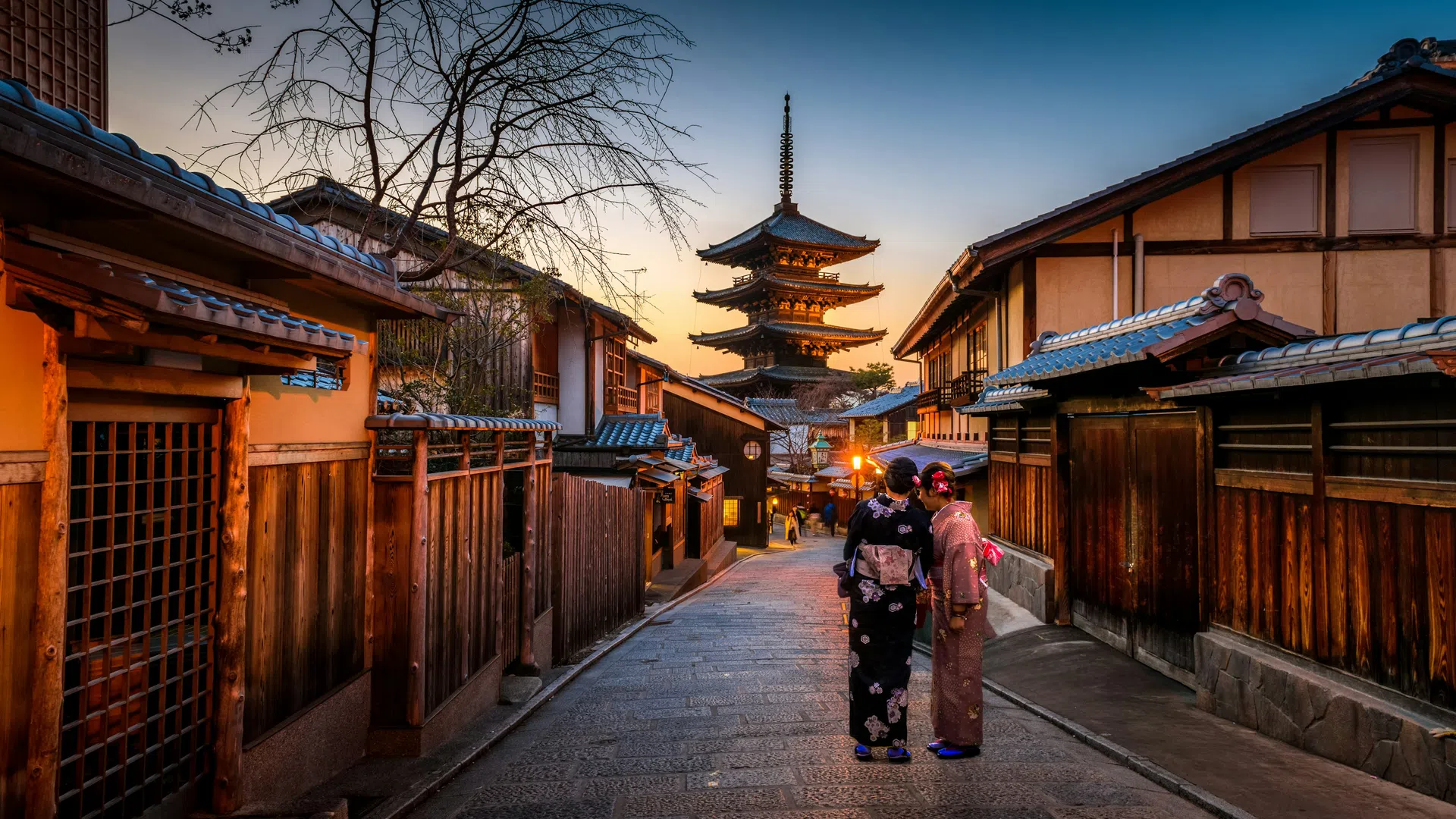 Two women in traditional kimonos walk through a historic street at sunset with a five-story pagoda in the background.