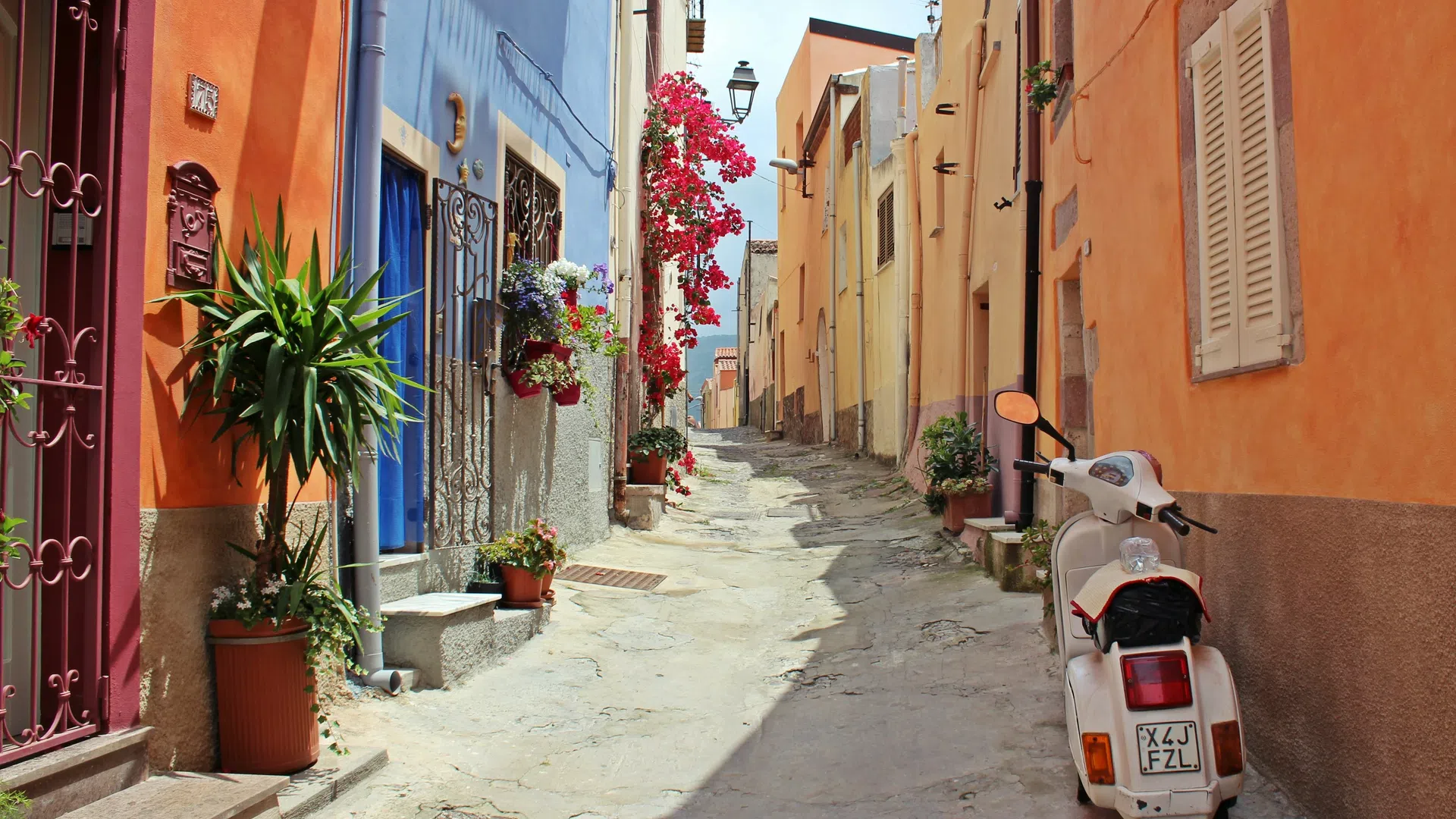 Narrow cobblestone street lined with colorful houses and potted plants with a white scooter parked on the side.