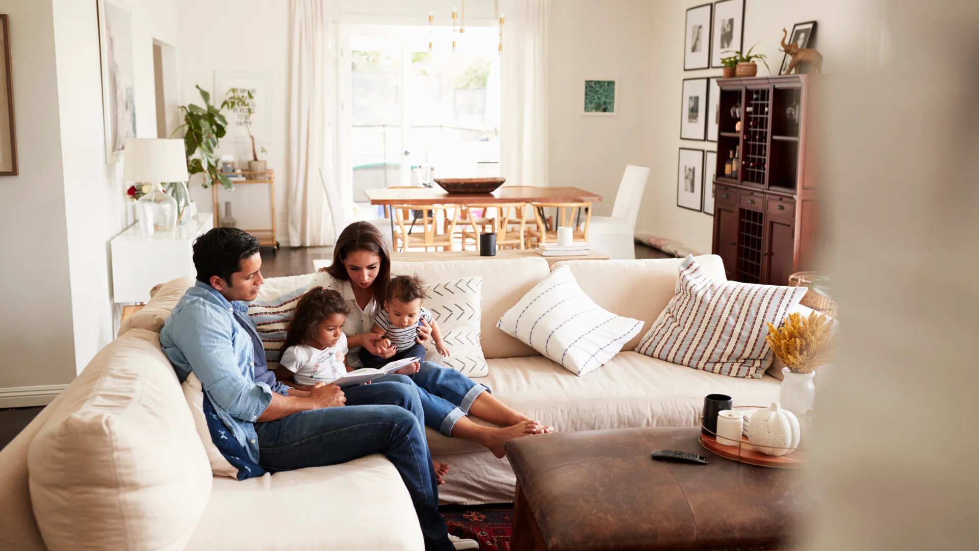 Family of four sitting on beige couch reading a book in a bright, cozy living room with modern decor