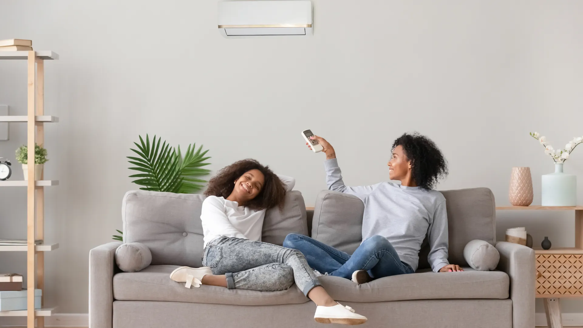 Two women relaxing on a gray couch in a modern living room with a remote control and air conditioner above