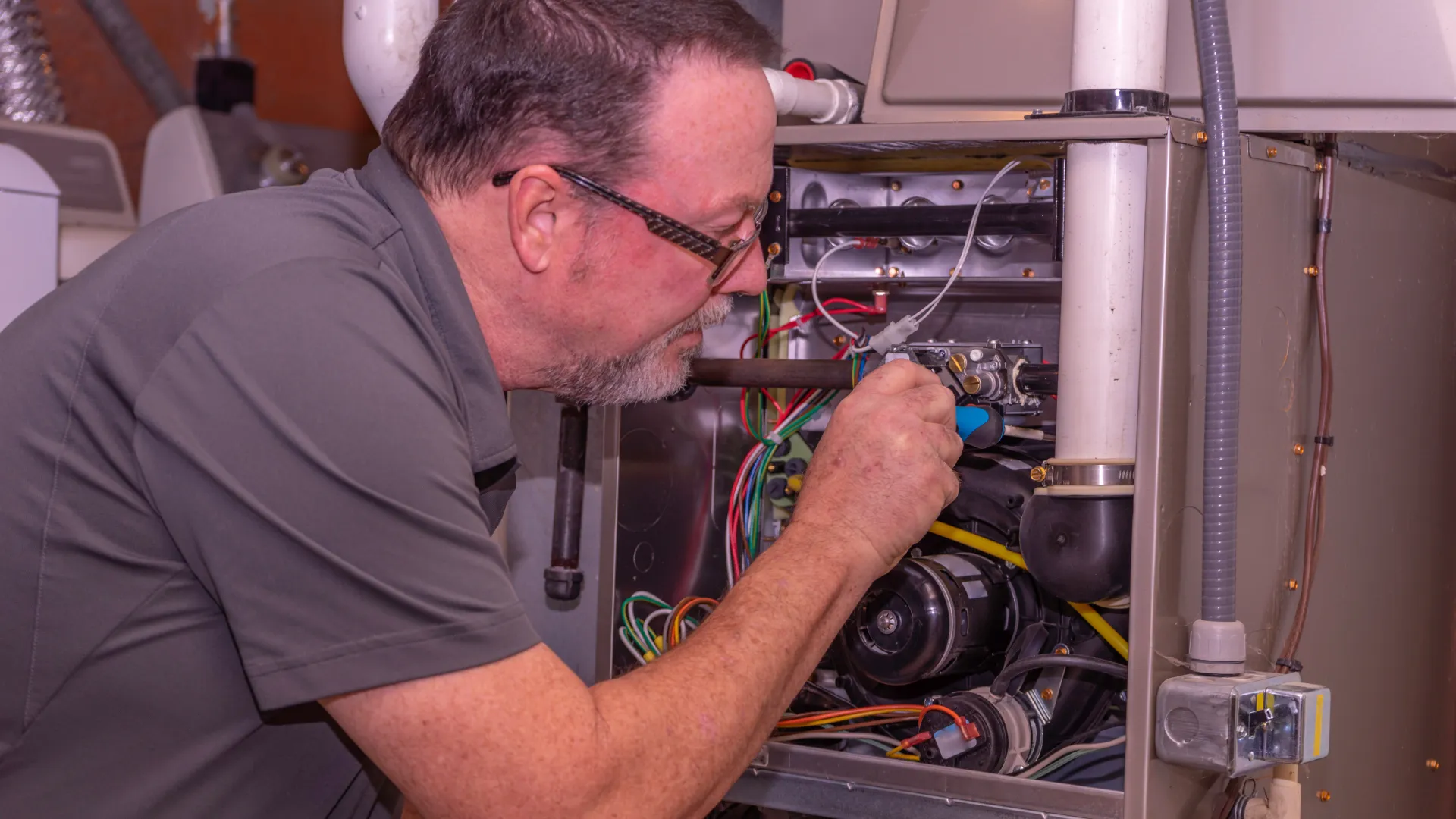 Technician repairing and inspecting a residential furnace with various wires and pipes in a utility room.