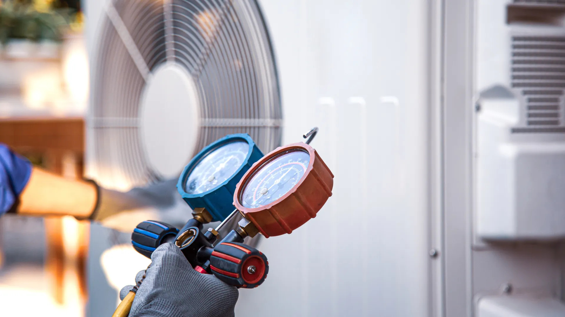 Technician's hand in glove holding HVAC manifold gauge set near outdoor air conditioning unit fan.