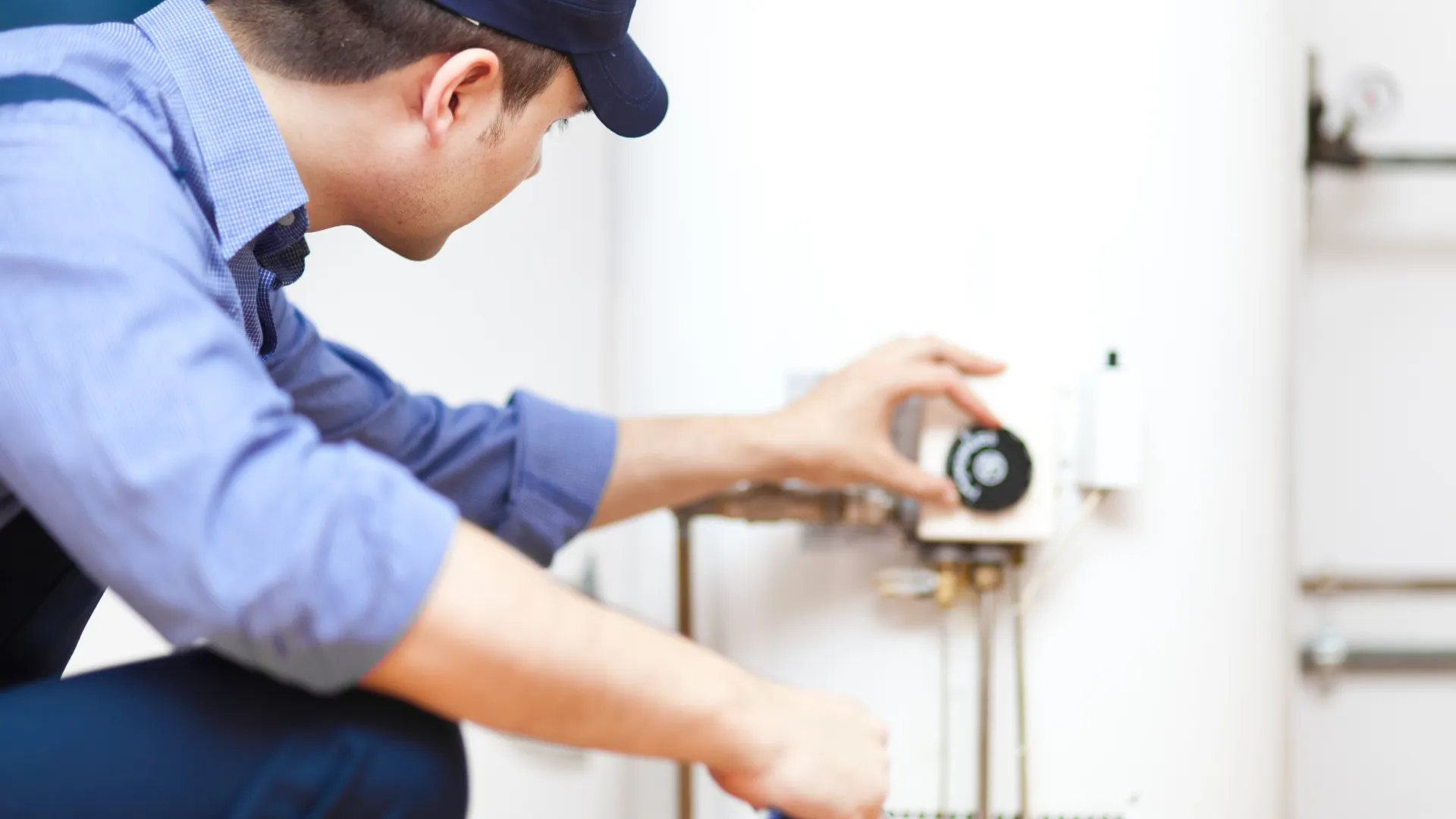 Technician adjusting controls on a white water heater in a maintenance setting wearing a blue uniform and cap