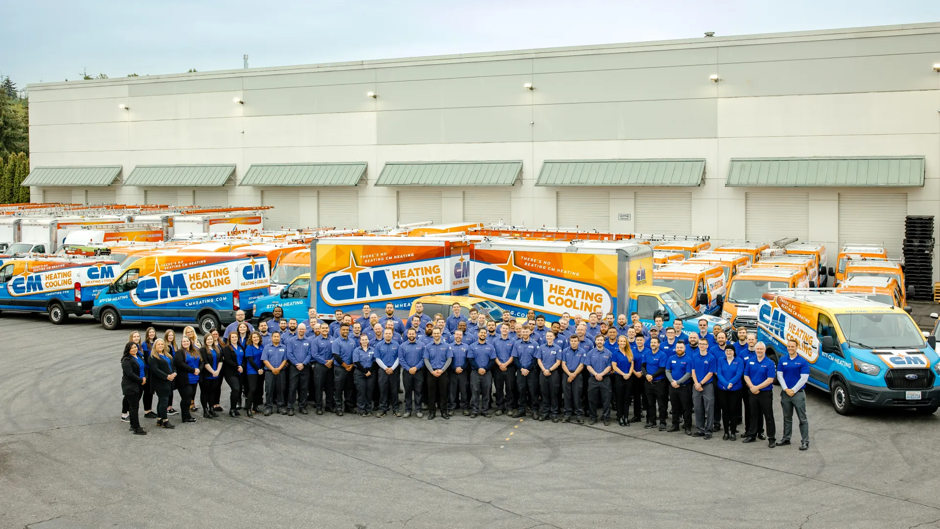 Large group of CM Heating Cooling employees posing in front of company service vans outside warehouse.