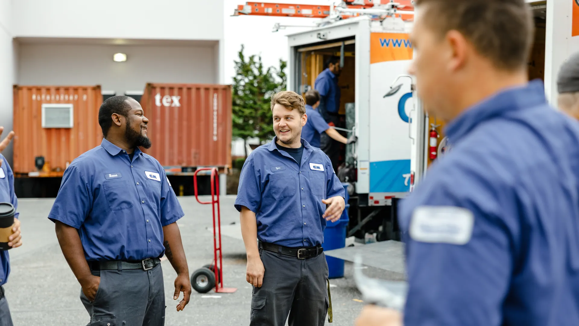 Group of uniformed technicians smiling and talking outside near work trucks and equipment on a sunny day