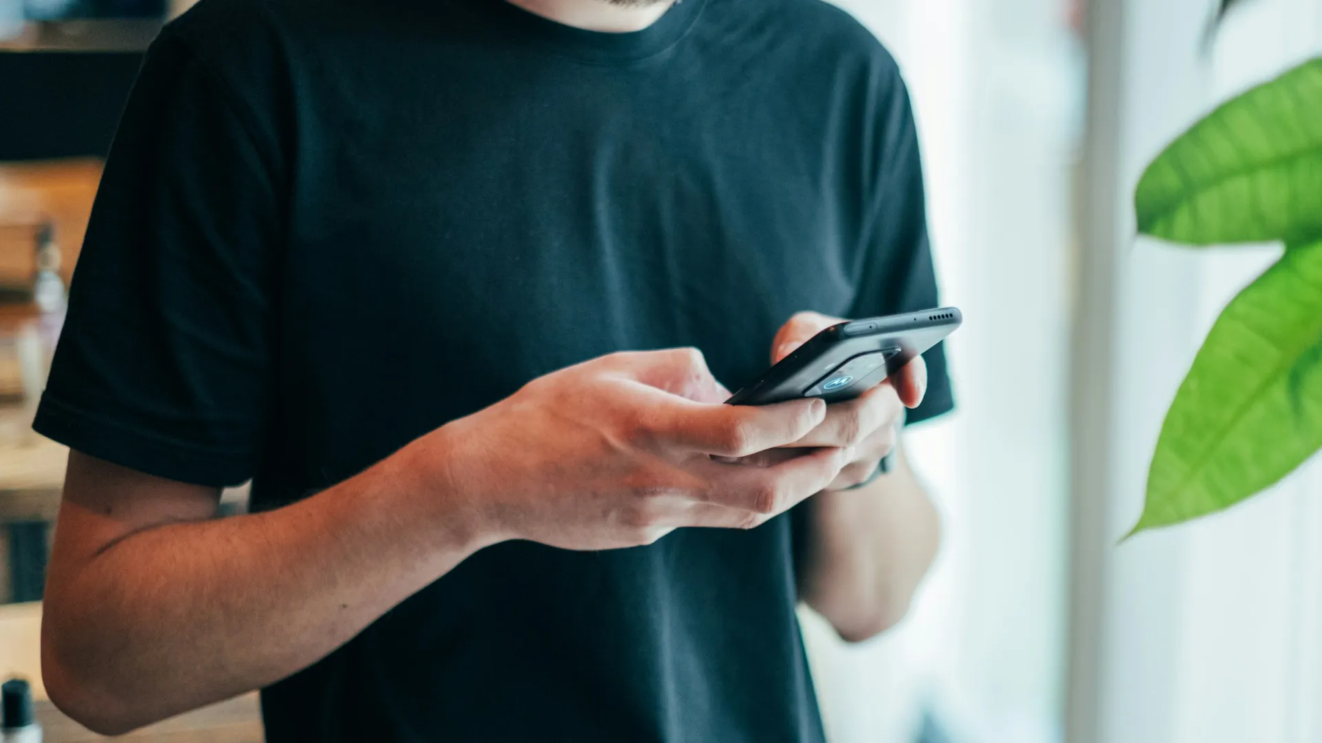 Man in black shirt using smartphone indoors near window with green plant leaves visible