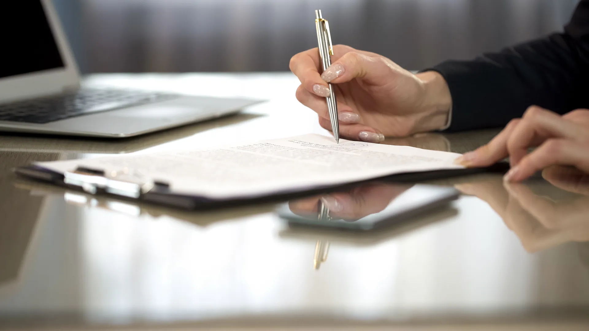 Person signing a document on a clipboard with a pen at a glass desk with a laptop nearby.