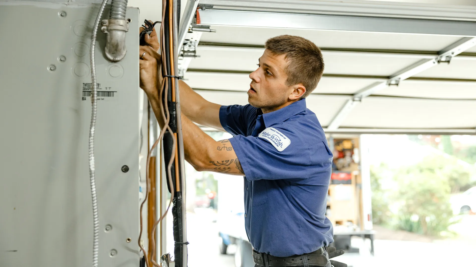 Electrician in blue uniform fixing wiring inside an electrical panel in a residential garage.