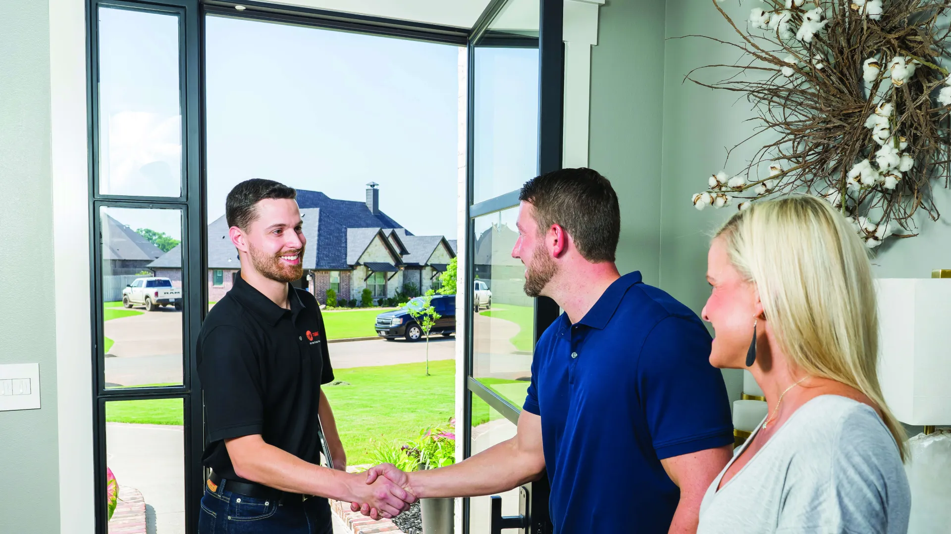 Man in black shaking hands with a smiling homeowner at the front door while a woman looks on indoors