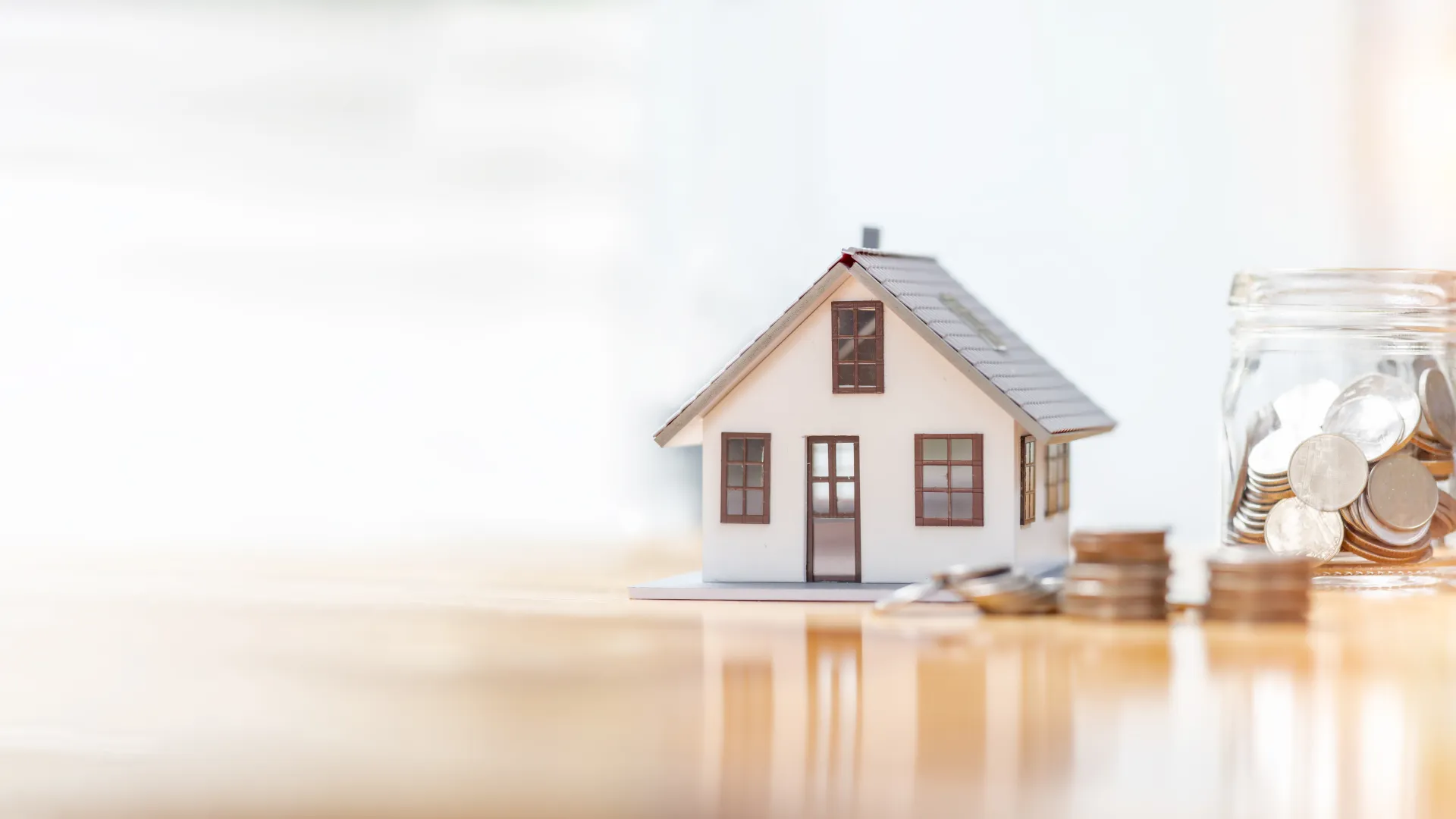 Miniature house model next to stacked coins and a jar filled with coins on a wooden surface.