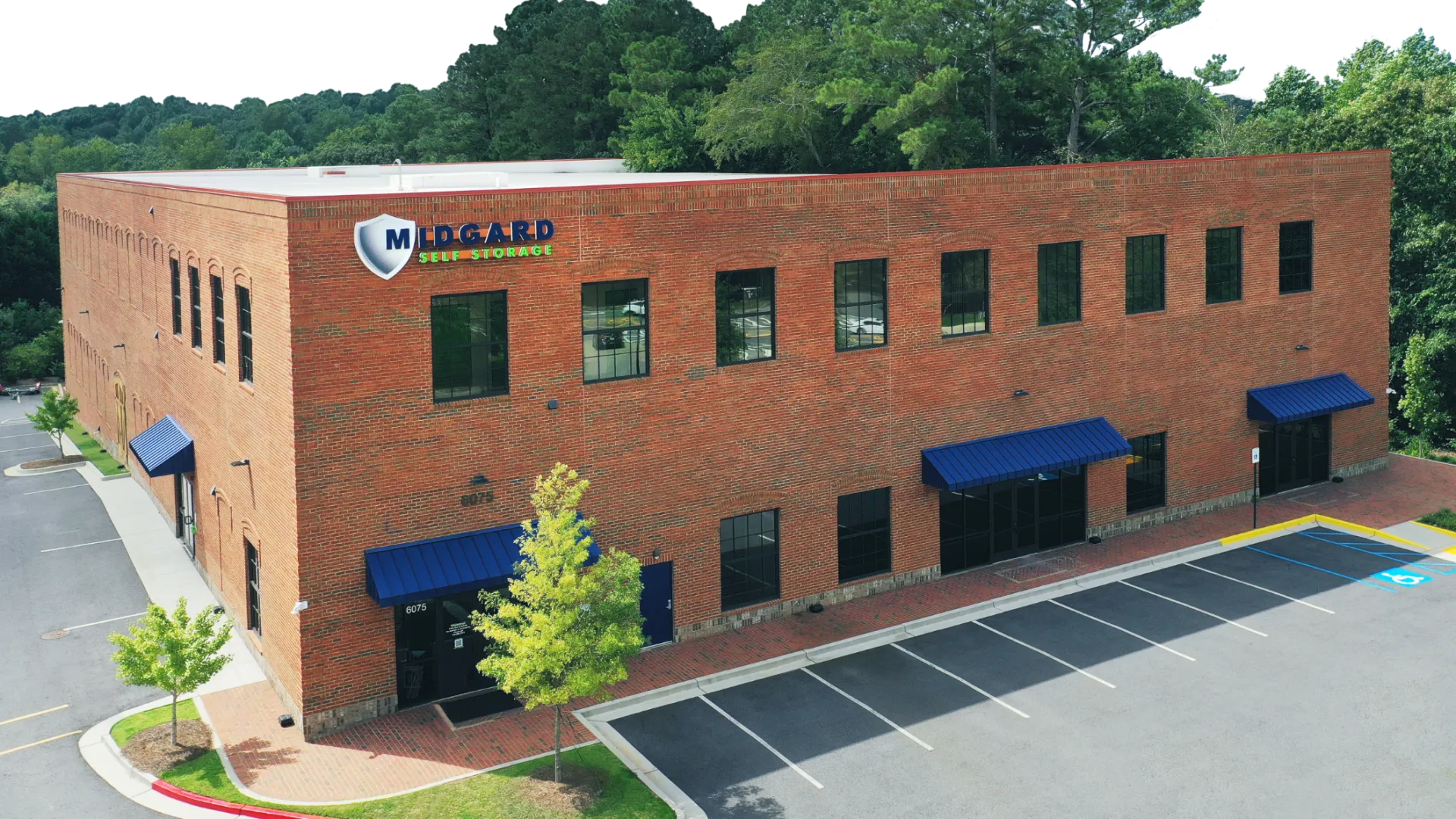 Two-story red brick building with blue awnings, parking lot, and Midgard Self Storage sign surrounded by greenery.
