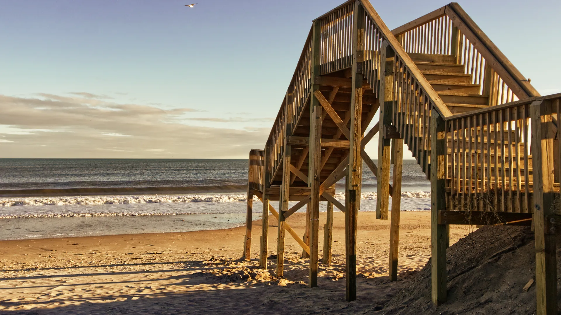 Wooden stairs leading down to a sandy beach with ocean waves and a bird flying in the sky at sunset.