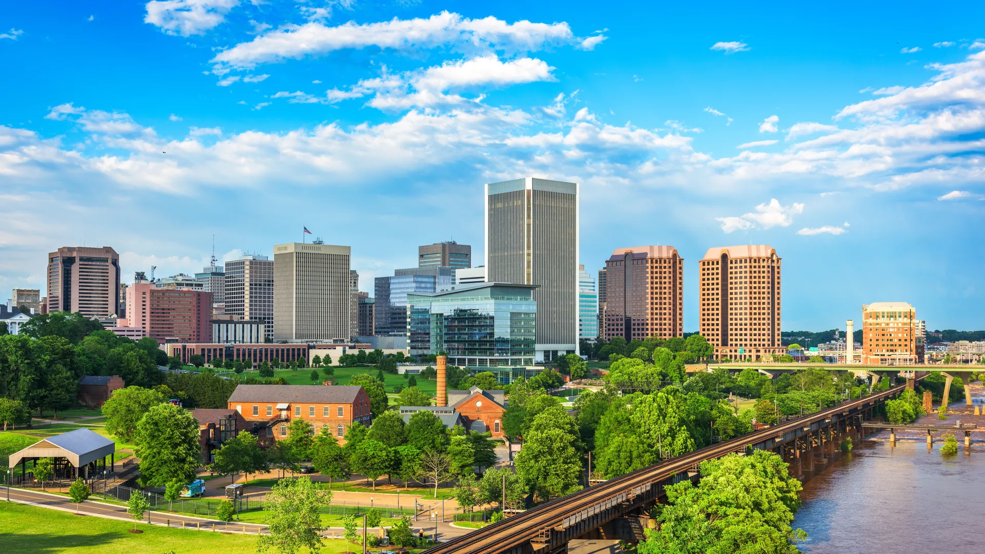 Richmond Virginia skyline with modern buildings, green parks, river, and railroad tracks under a partly cloudy sky.