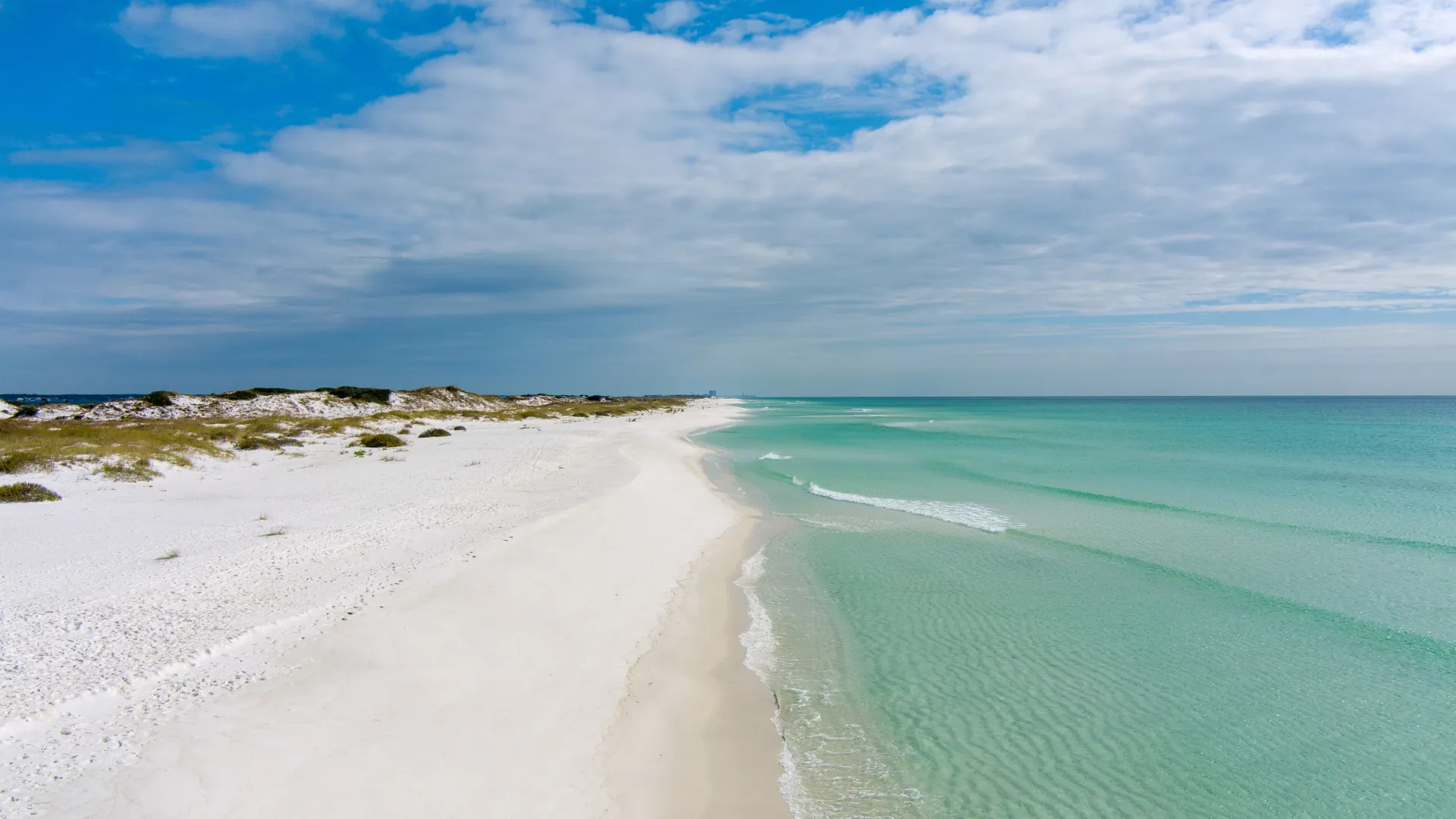 White sandy beach with clear turquoise water under a partly cloudy blue sky along a tranquil coastline.