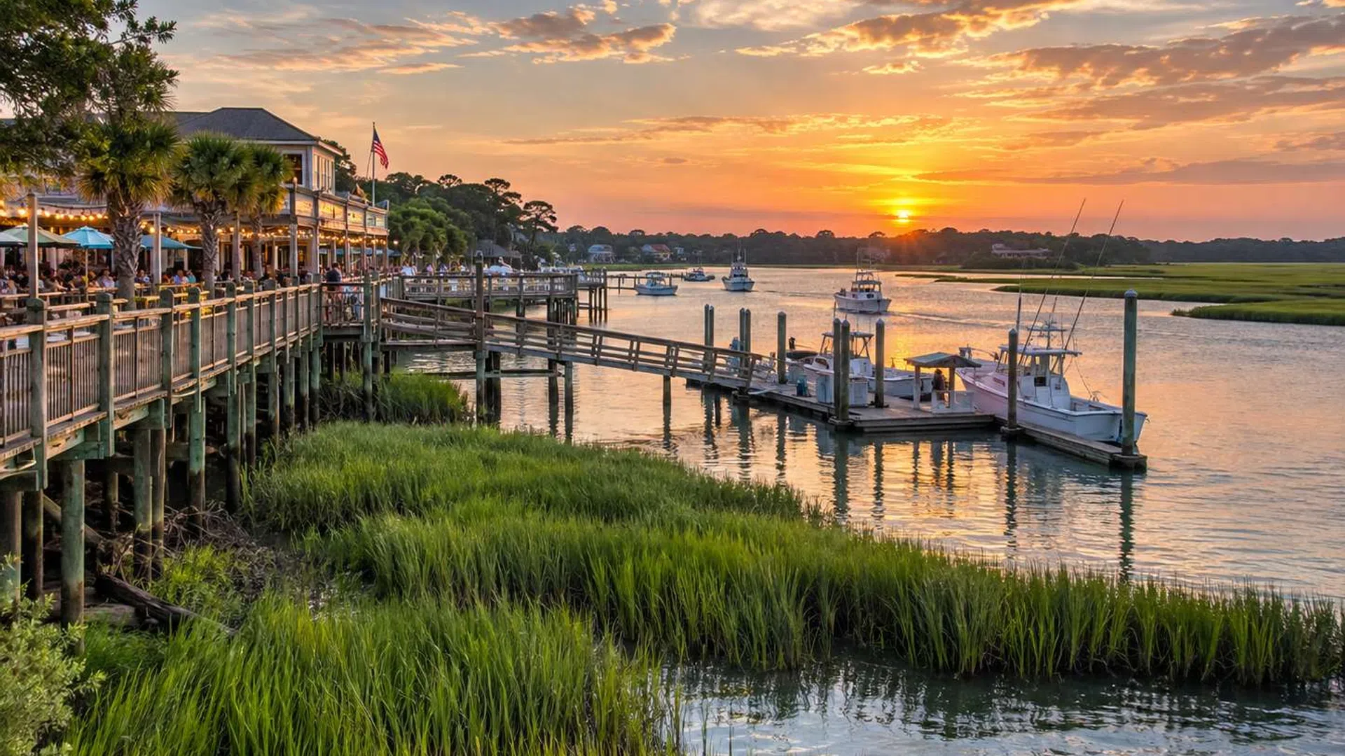 Sunset over a riverside dock with boats, grassy marshland, and a lively waterfront restaurant with outdoor seating.