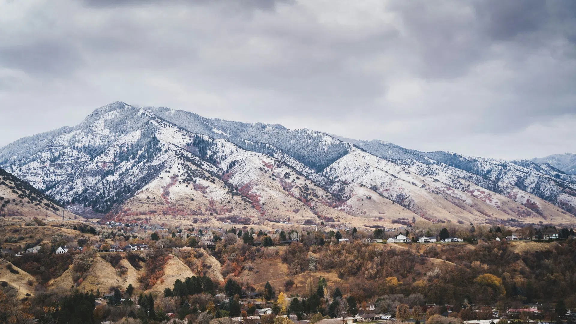 Mountain range with light snow under cloudy sky overlooking autumn-colored forest and scattered houses.