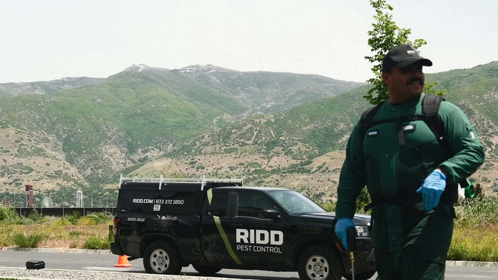 Pest control technician in green uniform with sprayer near RIDD Pest Control truck in grassy suburban area.