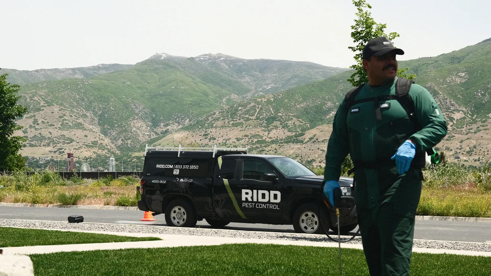 Pest control worker in green uniform spraying lawn with RIDO Pest Control truck parked nearby in a suburban area.