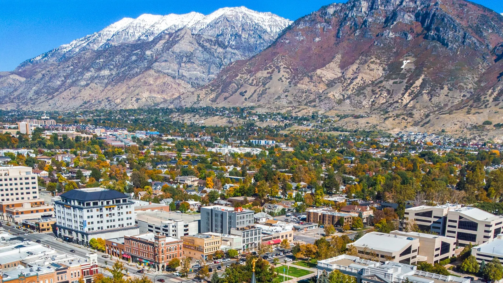 Panoramic view of a city with buildings, trees, and snow-capped mountains under a blue sky.
