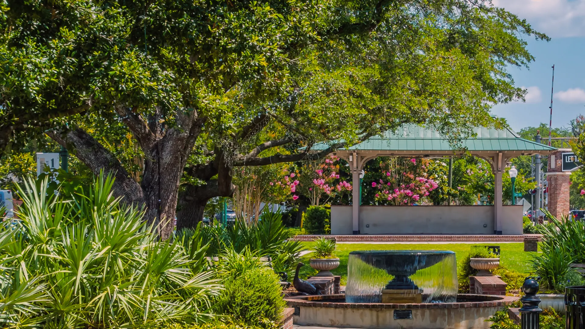 Sunny park scene with lush green trees, a flowing fountain, and a gazebo surrounded by vibrant flowers.