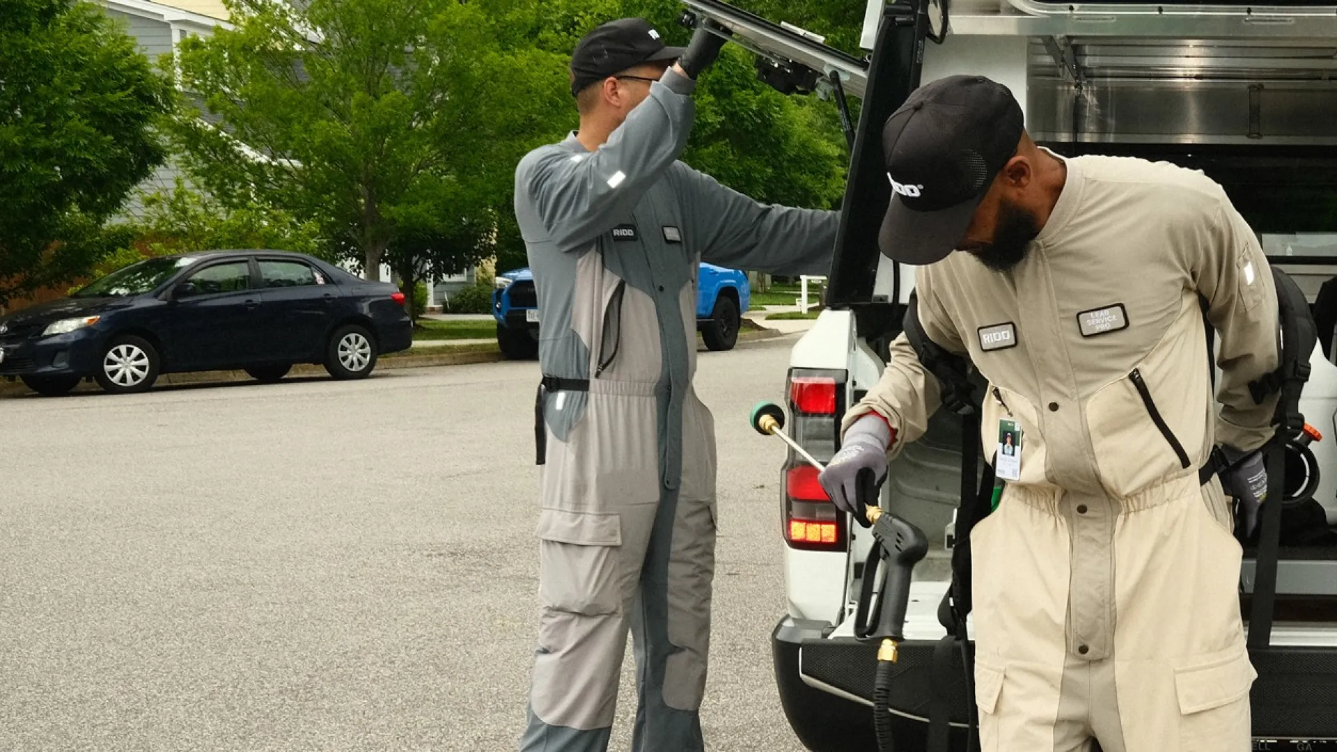Two pest control workers preparing equipment at the back of a service truck in a suburban neighborhood.