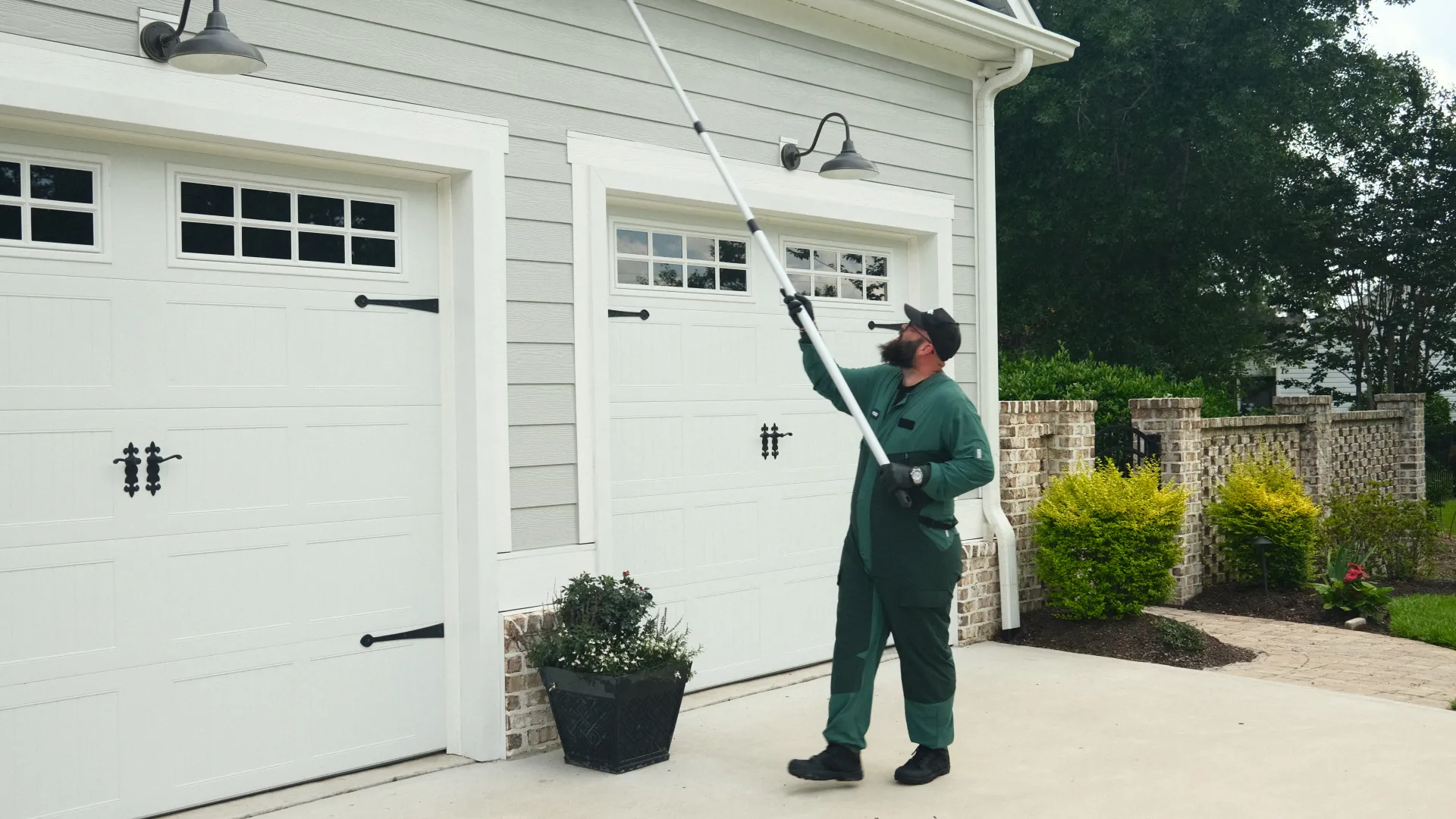 Man in green coveralls using a pole tool to clean outdoor light fixtures on a house exterior near garage doors.