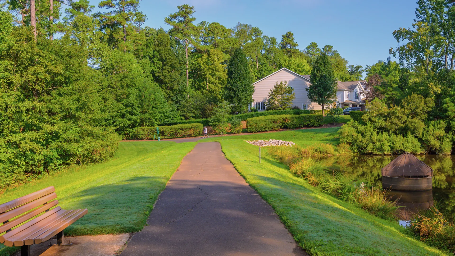 Paved path in a green park with a bench, trees, a pond, and houses in the background under clear blue sky